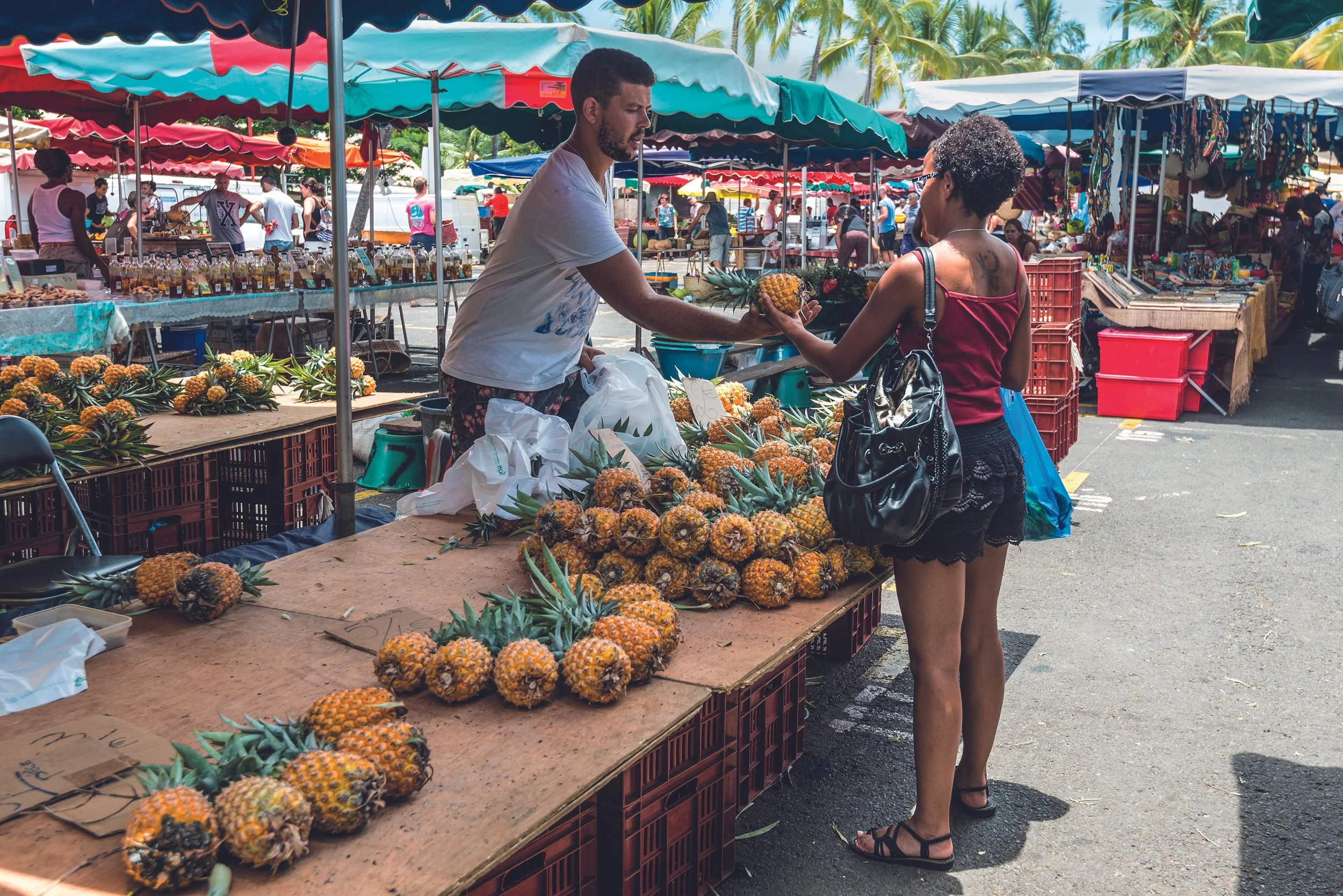 Une vendeuse vend des ananas à un client dans un marché en plein air avec des stands colorés et des palmiers en arrière-plan.