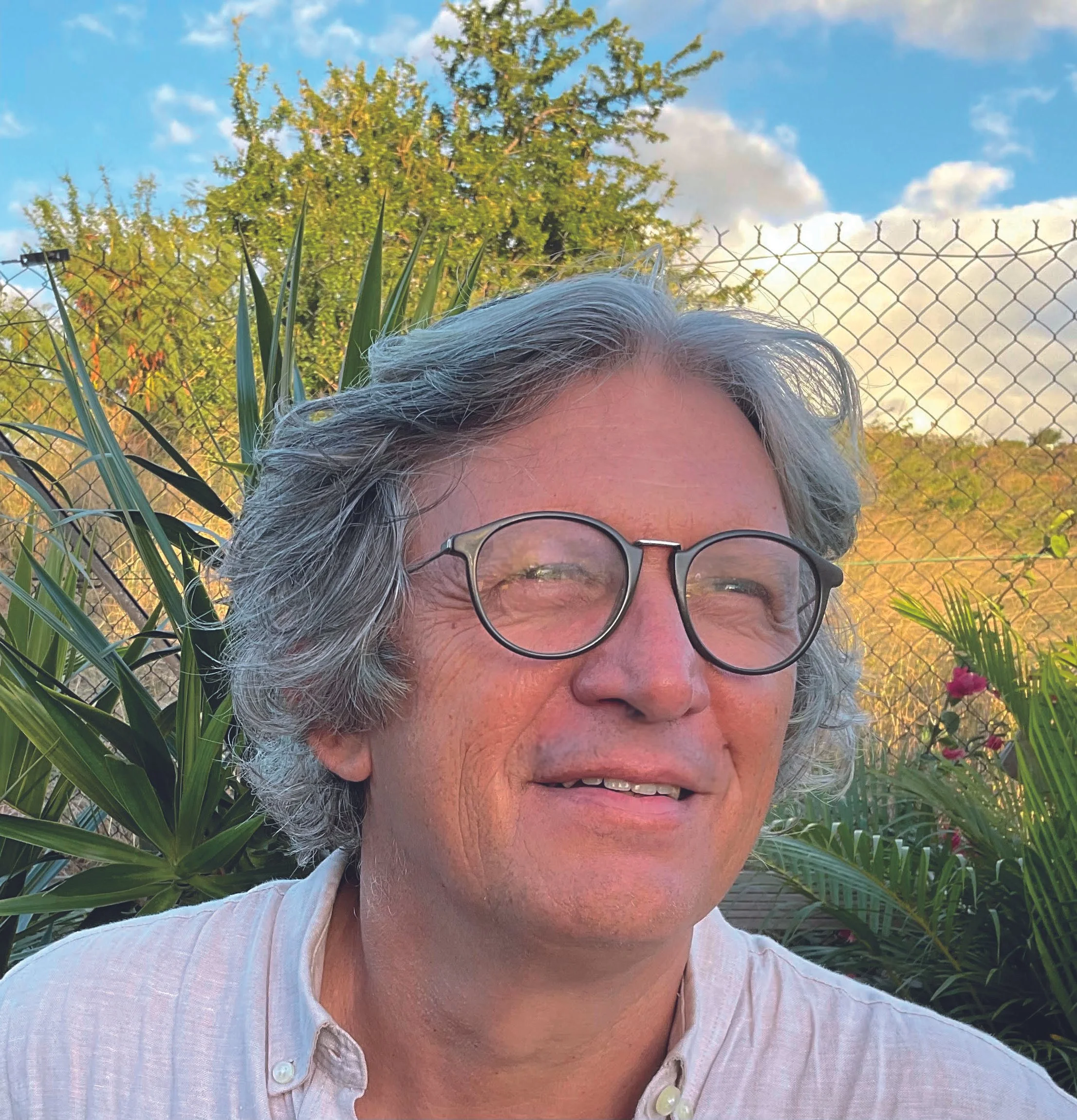 Un homme avec des lunettes, souriant, devant un jardin avec des plantes vertes et un ciel partiellement nuageux.