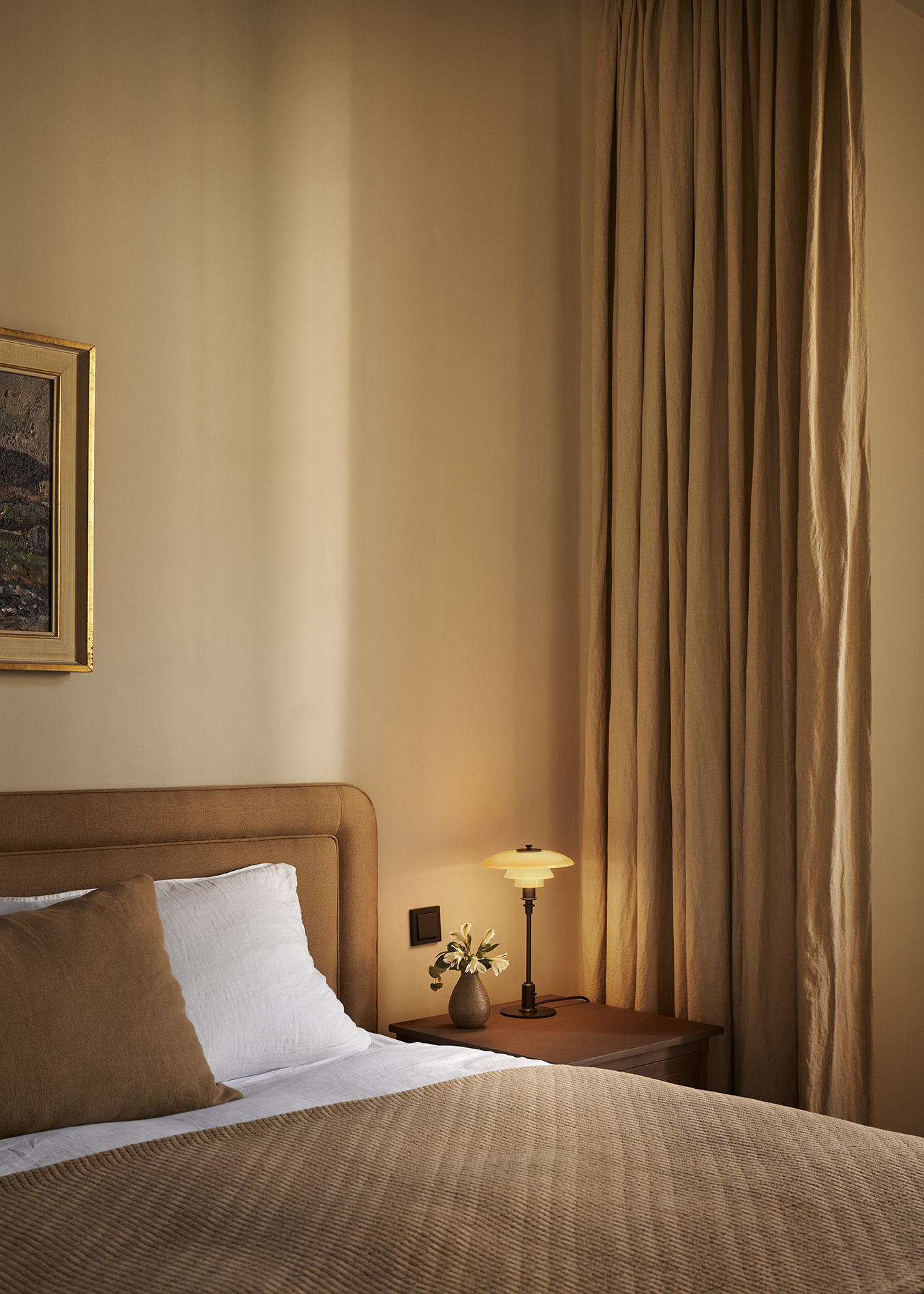 Cozy bedroom with beige bedding, elegant curtains, a bedside table with a PH 2/1 Limited Edition Table lamp, a small vase with flowers, and a framed painting on the wall. Staged by Swedish Design Duo DuRietz Design, campaign work for Louis Poulsen.
