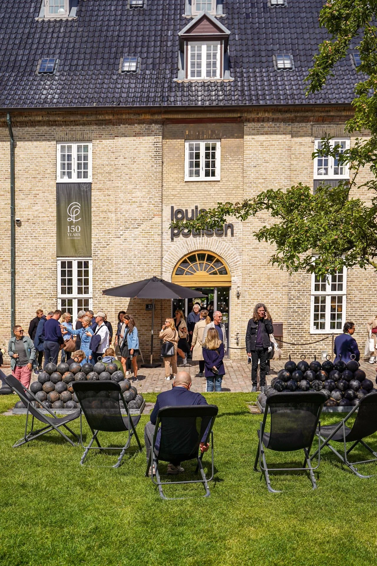 Group of people outside a historic building with "louis poulsen" sign, on a sunny day, with modern outdoor seating and spherical sculptures. Heart of Light 3daysofdesign Copenhagen, by Frederik Nystrup-Larsen and Oliver Sundquist. PH Artichoke, Louis