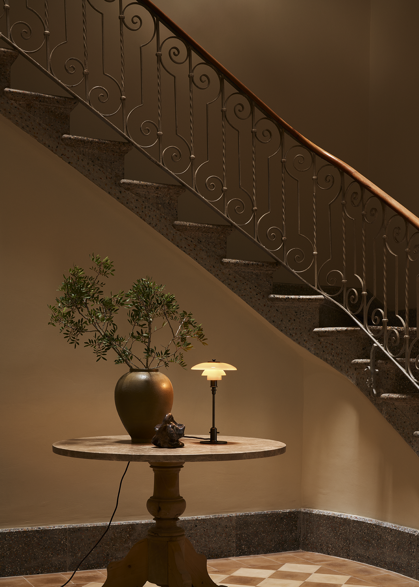 Elegant stairway with ornate metal railing, wooden table, vase with greenery, and stylish PH 2/1 Limited Edition table lamp. Styled by swedish design duo DuRietz Design.