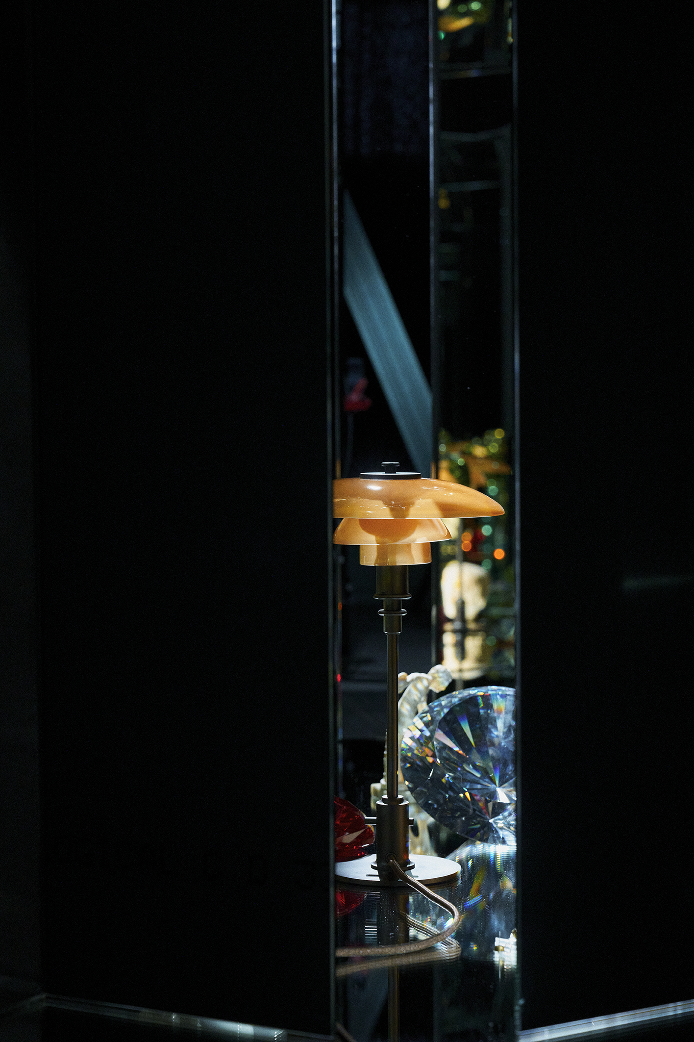 A PH 2/1 Table lamp, Limited Edition Dusty Terracotta, with a layered design is placed inside a dark display case, reflecting light on a nearby large, faceted crystal object. Staged by Danish Artist Esben Weile Kjær, campaign work for Louis Poulsen.