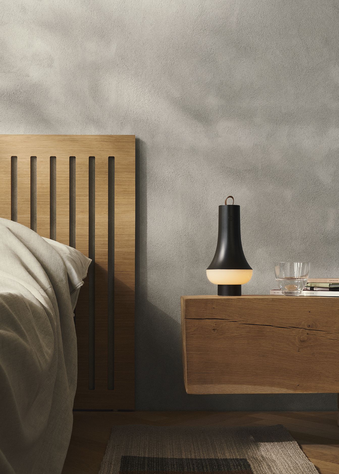 Modern bedroom scene with wooden headboard, floating nightstand, stylish Tomoshi lamp by nendo Oki Sato, glass cup, and books against a textured gray wall.