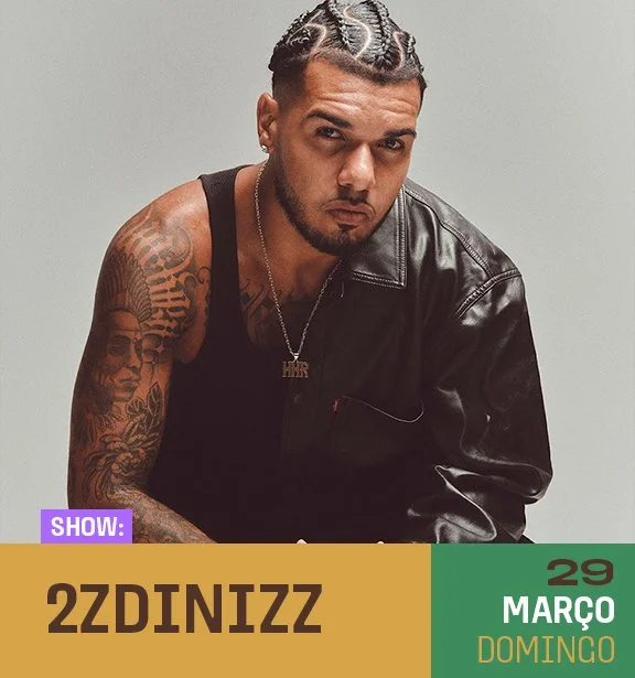 Homem com cabelo trançado, tatuagens nos braços, usando jaqueta de couro preto e colar com pingente, posando contra fundo cinza