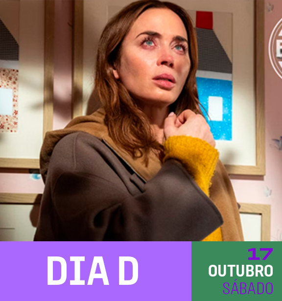 Mulher com cabelo castanho e olhos claros, usando um casaco cinza e uma blusa amarela, olhando para cima com expressão pensativa, em um ambiente com quadros coloridos na parede ao fundo.