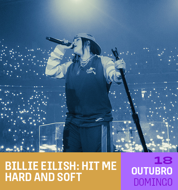 Uma pessoa no palco segurando um microfone e um boom pole, durante um show ou apresentação, com uma grande plateia de luzes de celulares acesos ao fundo, sob iluminação de palco azul com o céu escuro.