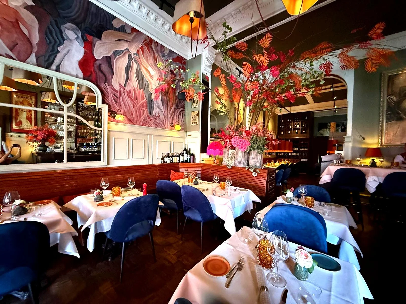 Elegantes Restaurant mit weiß gedeckten Tischen, blauen Stühlen, einer großen Blumenampel mit pinken und orangefarbenen Blumen, Wandgemälde und goldenen Lampen.