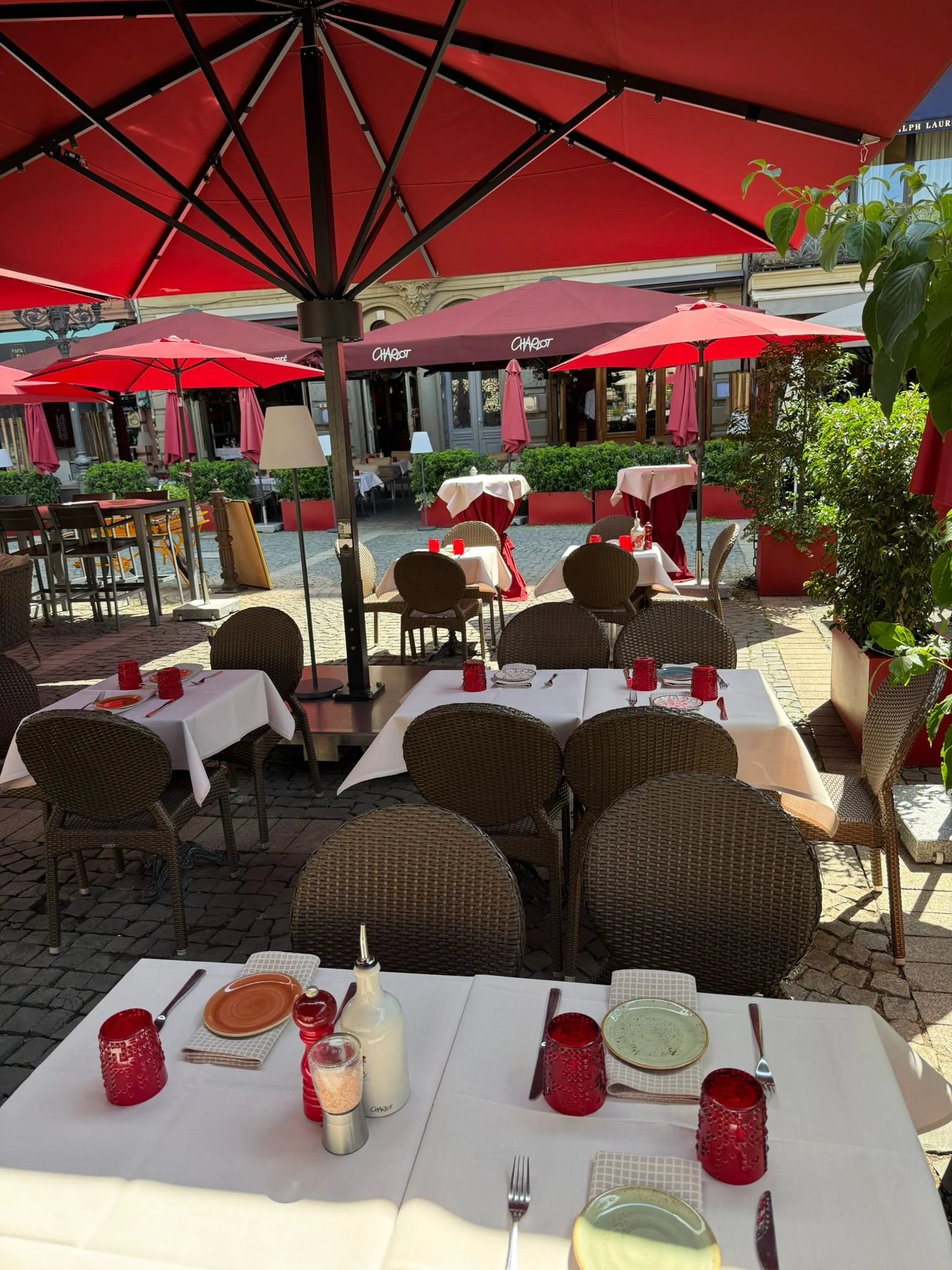 Außenbereich mit Tischen, Stühlen und großen roten Sonnenschirmen, Untergrund ist Kopfsteinpflaster, im Hintergrund O/C Operncafé. Es ist sonnig und es gibt viel Grünpflanzen.