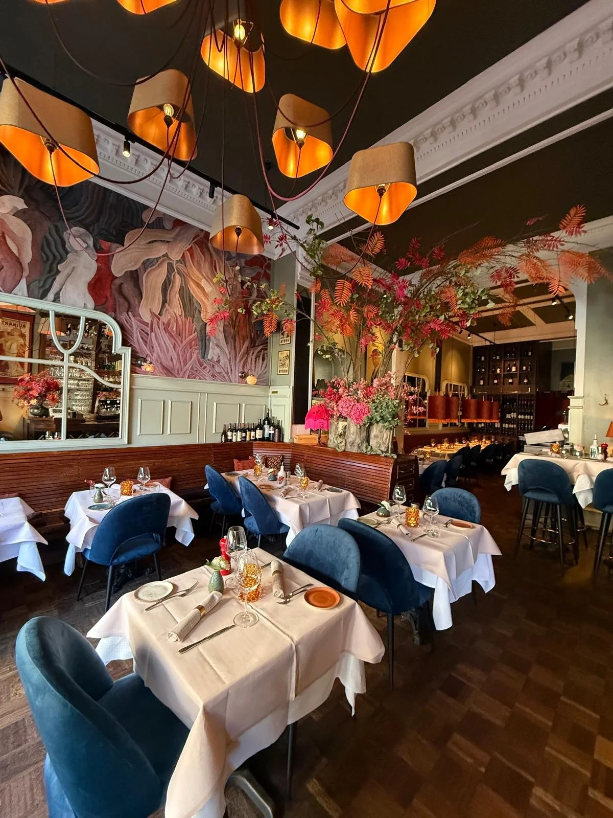 Elegantes Restaurant mit weißen Tischtüchern, blauen Sesseln und großen orangefarbenen Lampenschirmen, lebendige Blumendekoration und Wandbild im Hintergrund.