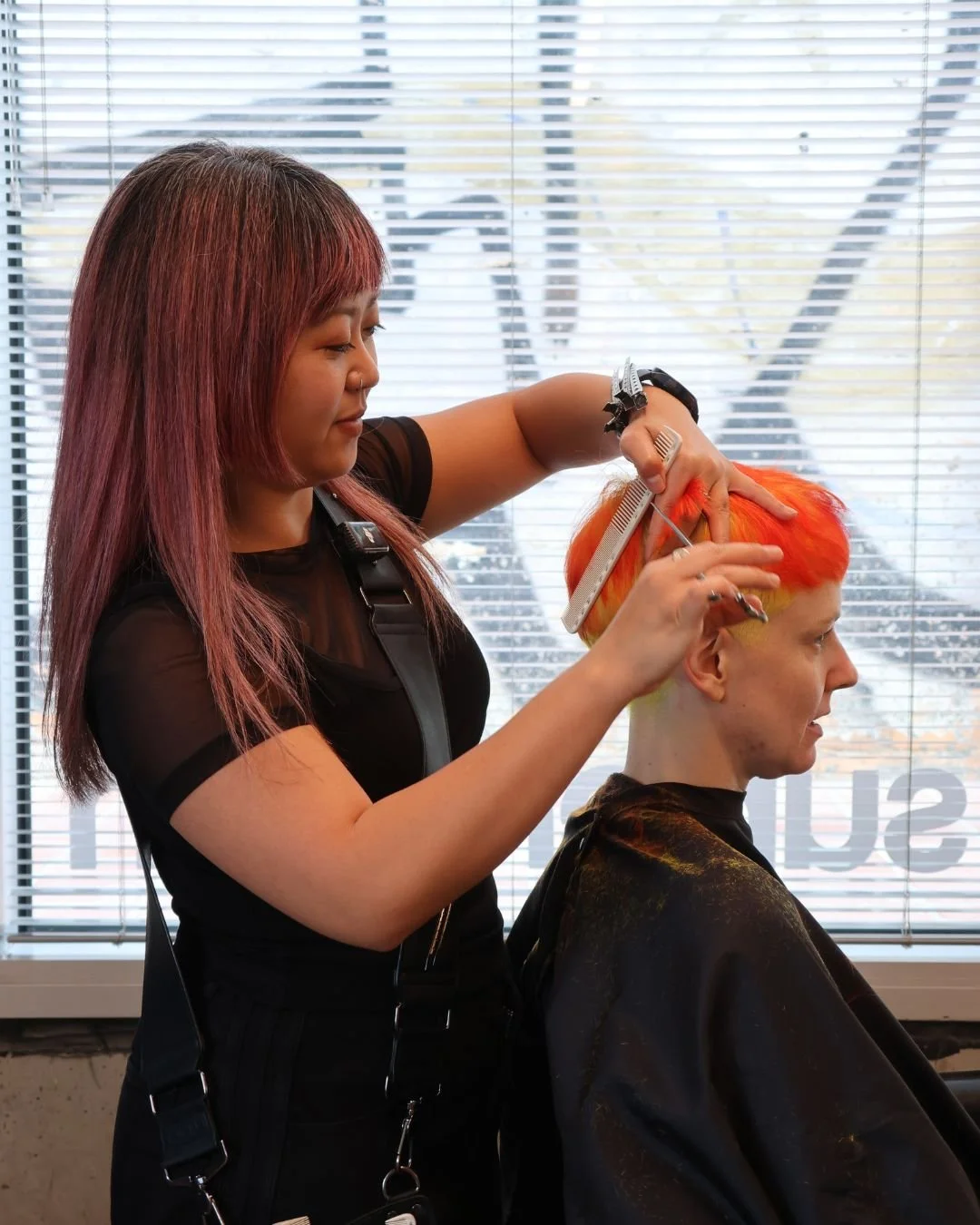 A woman with vibrant orange and yellow hair is getting a haircut by a stylist with long, reddish-brown hair. The stylist is using scissors and a comb, standing in front of a window with blinds in a salon.