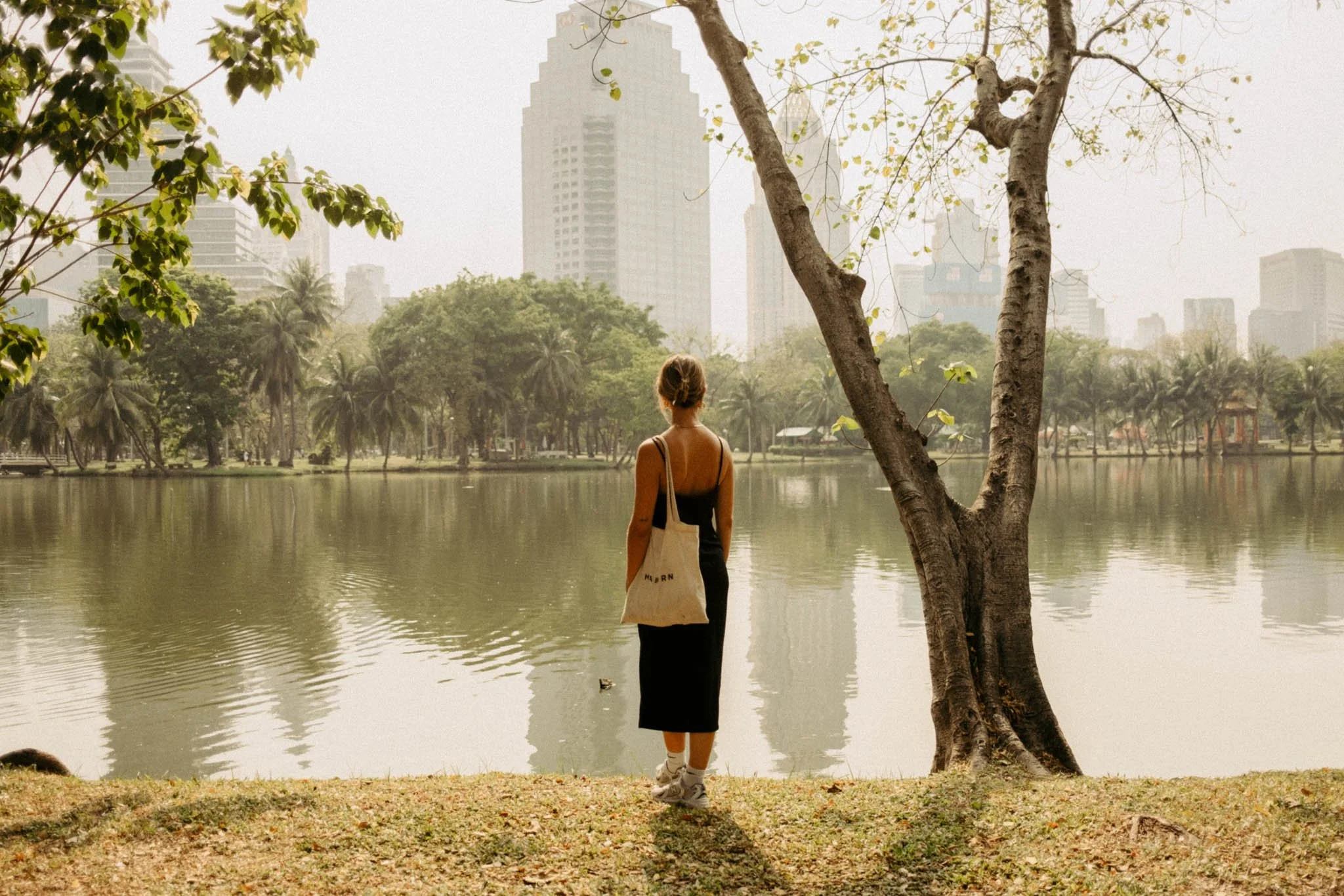 Une femme debout au bord d'un lac dans un parc urbain, avec des gratte-ciel en arrière-plan, en automne ou en fin d'après-midi.
