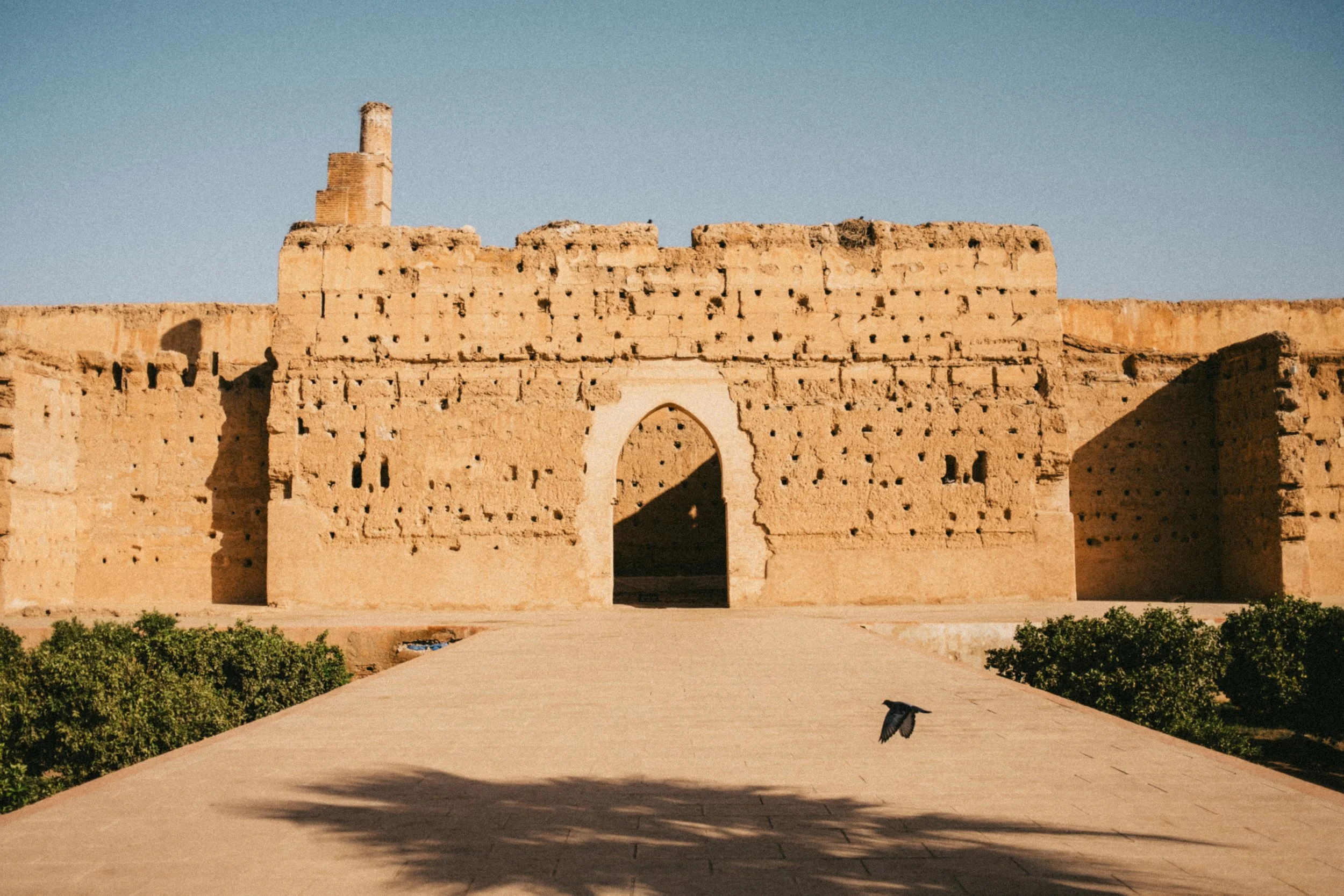 Structure en pierre ancienne avec une arche centrale et un mur partiellement en ruines, sous un ciel clair, avec un chemin en avant et des arbustes au sol.