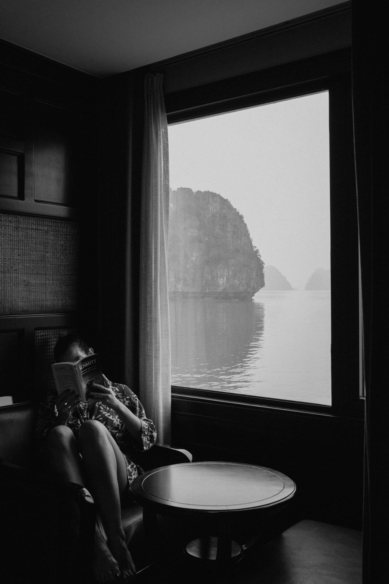 Person lisant un livre dans une pièce, à côté d'une grande fenêtre qui montre un paysage de falaises et d'eau en arrière-plan, en noir et blanc.