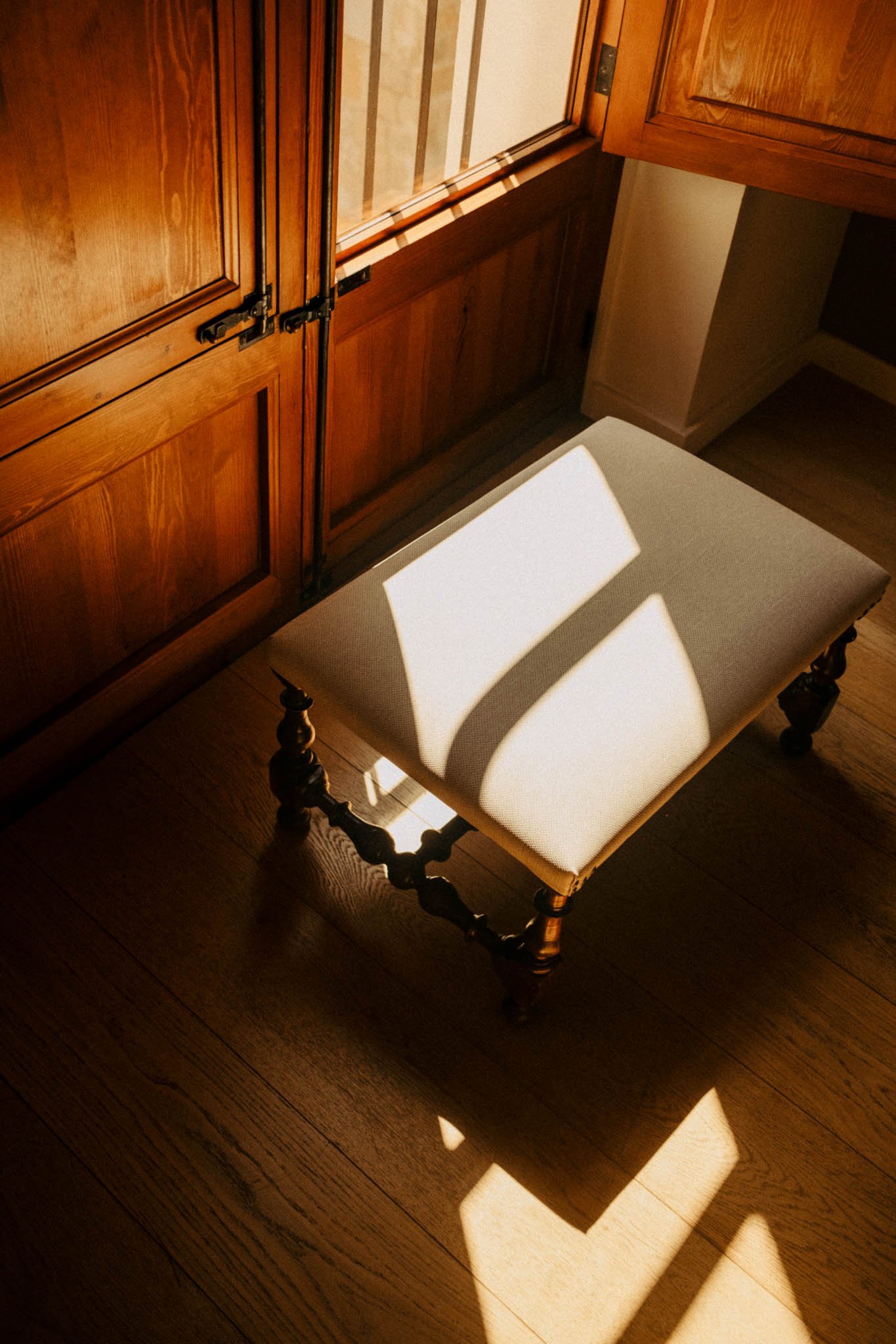 Une chaise avec un coussin beige sous la lumière du soleil, dans une pièce avec un sol en bois et des meubles en bois foncé.