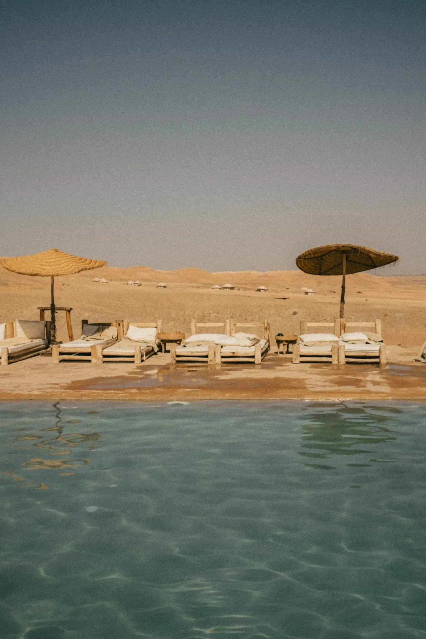 Chaises longues en bois avec des coussins, ombragées par des parasols en paille, au bord d'une piscine dans un désert, avec un ciel clair et des dunes en arrière-plan.