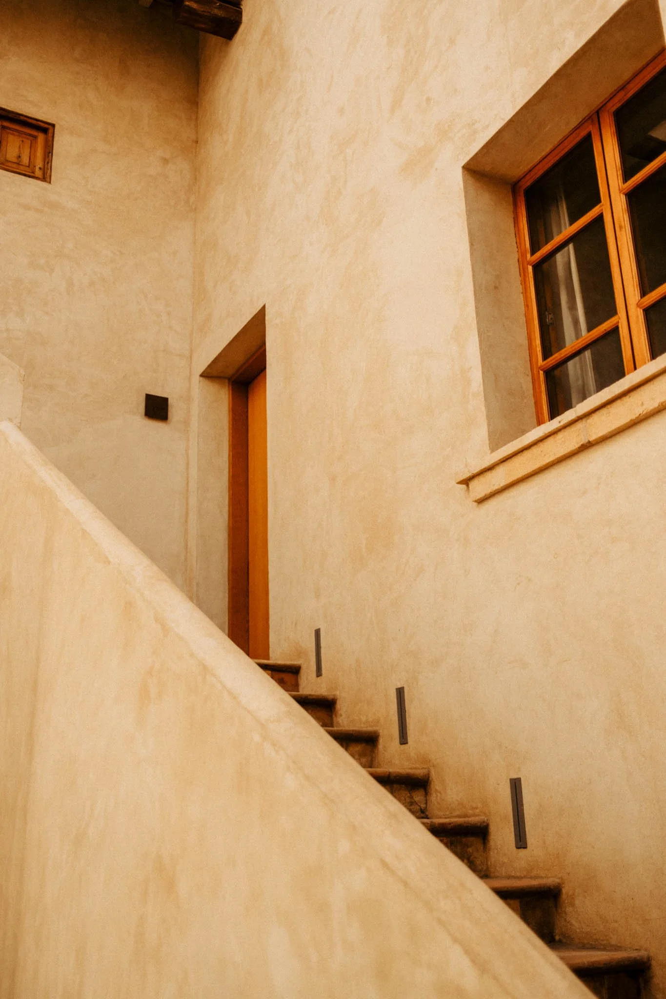 Vue d'un intérieur avec un escalier en pierre, une fenêtre à cadres en bois, et un mur en plâtre beige au style rustique.