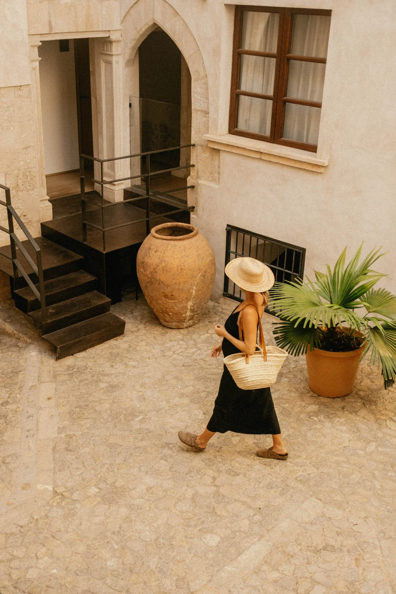 Femme portant un chapeau de paille noir, avec un sac en osier, marchant dans une cour pavée intérieure, avec une grande poterie, une plantation de palmier, et une fenêtre en bois, dans un style méditerranéen.