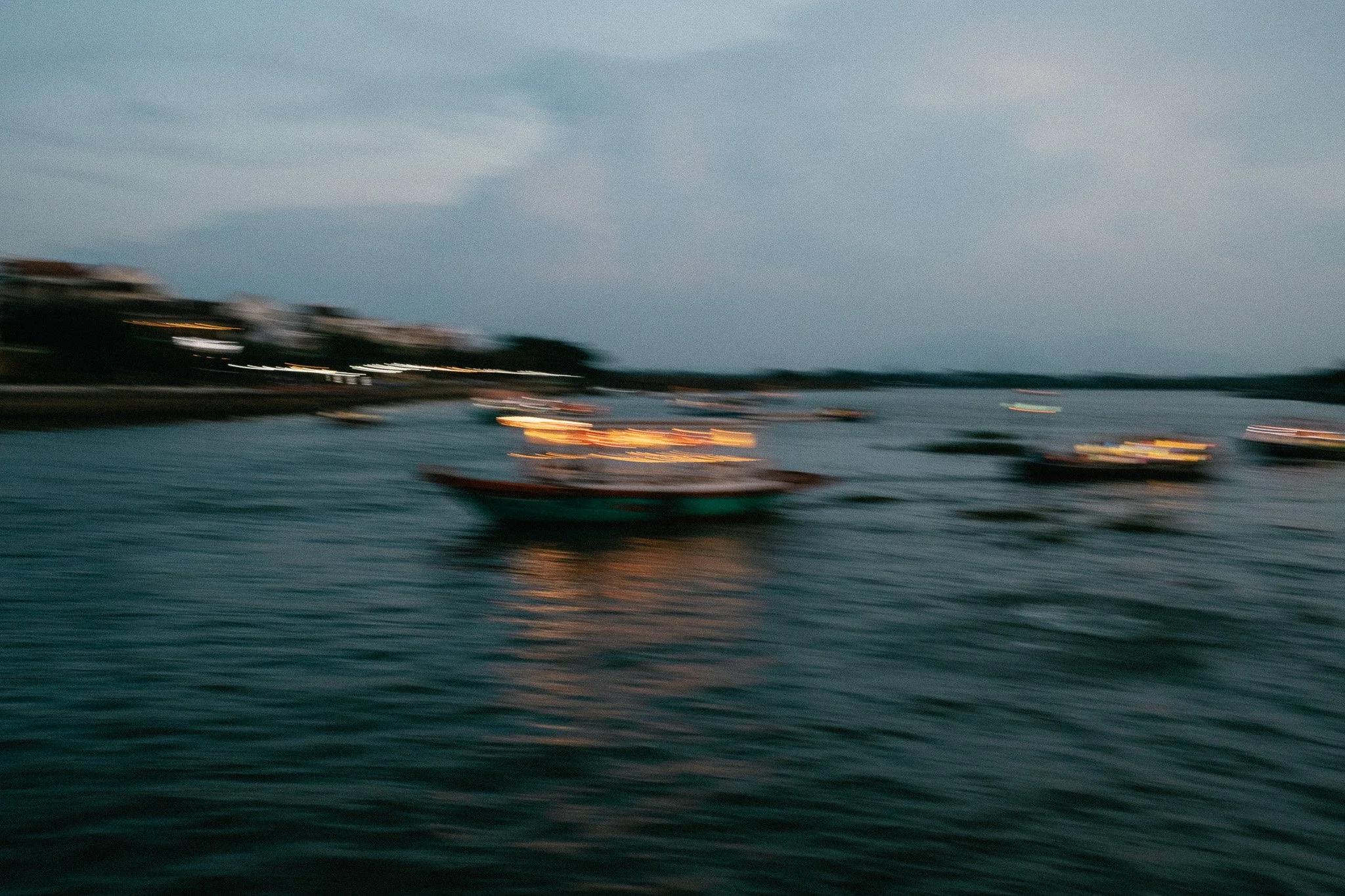 Bateau en mouvement sur un fleuve ou une mer, avec des lumières et bâtiments flous en arrière-plan lors d'un crépuscule ou au coucher du soleil.
