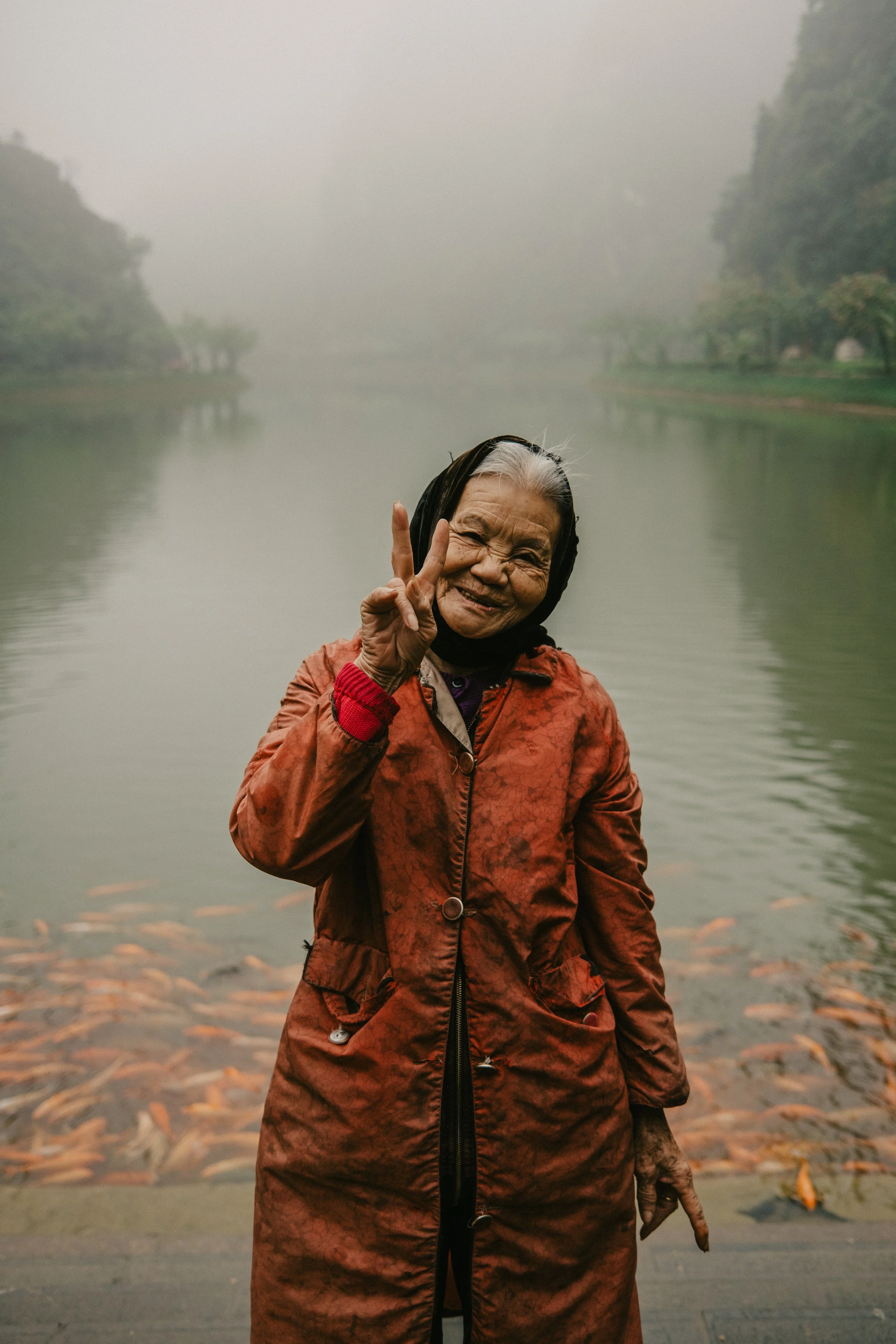 Une femme âgée souriante, portant un manteau rouge, fait un signe de paix avec ses doigts devant un étang avec des poissons, dans un environnement brumeux et naturel.