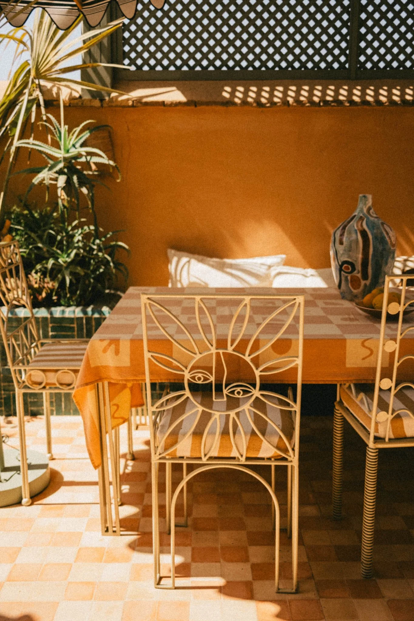 Terrasse extérieure avec une table, des chaises en fer blanc avec motifs floraux, un vase décoratif en céramique et des plantes vertes, sous une lumière naturelle