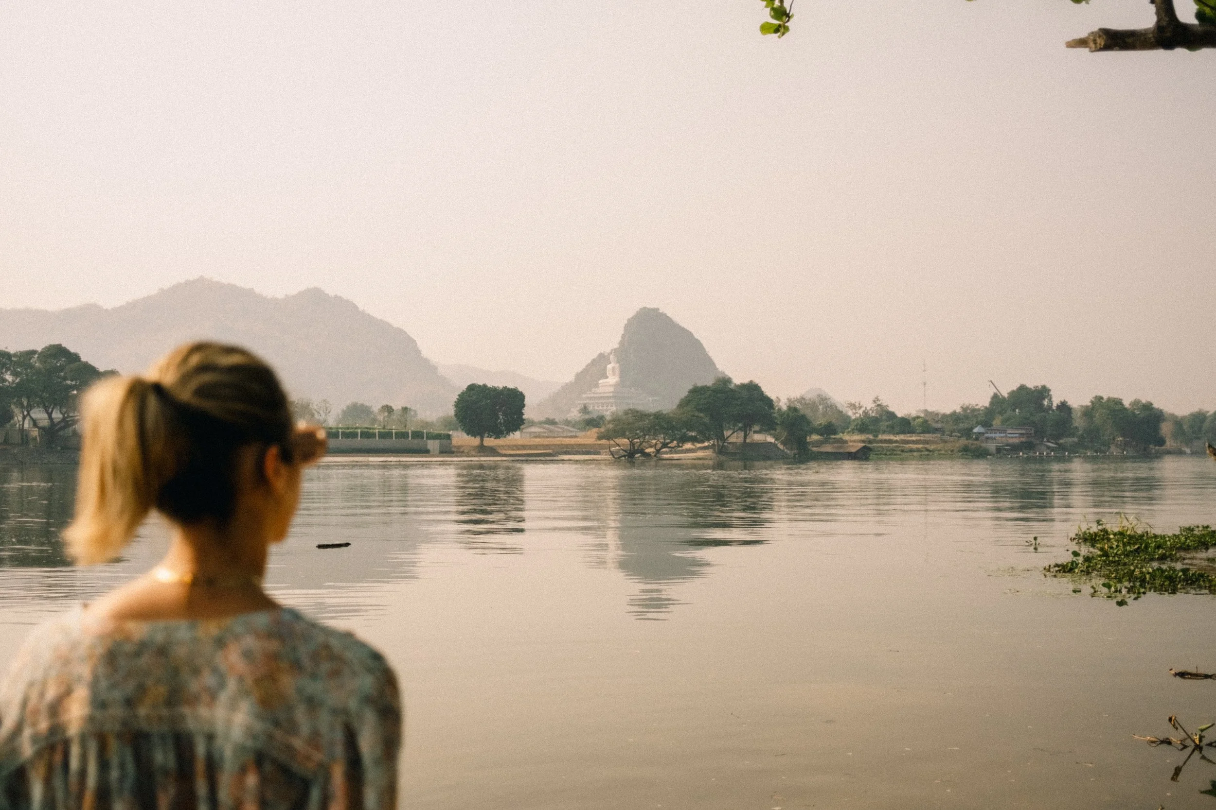 Une femme vue de dos regarde un fleuve avec une pagode et des montagnes en arrière-plan.