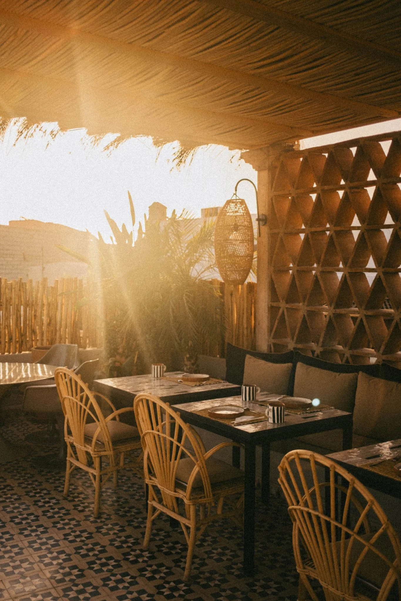 Terrasse de restaurant en plein soleil avec tables, chaises en bois, décor en bambou et lampes en rotin.