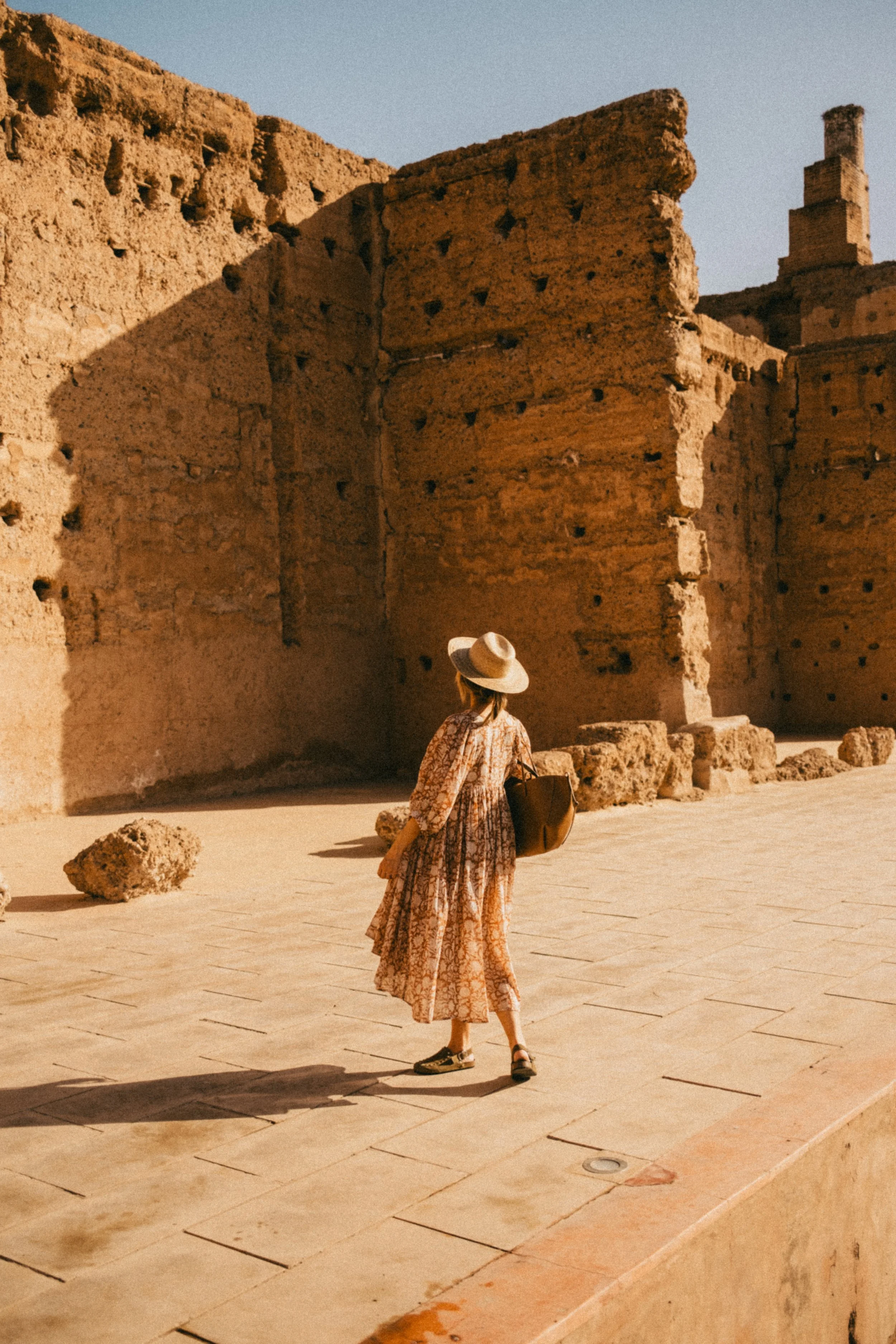 Une femme portant un chapeau, une robe longue à motifs et un sac, se promène devant un ancien mur de ruines en pierre dans un paysage ensoleillé.