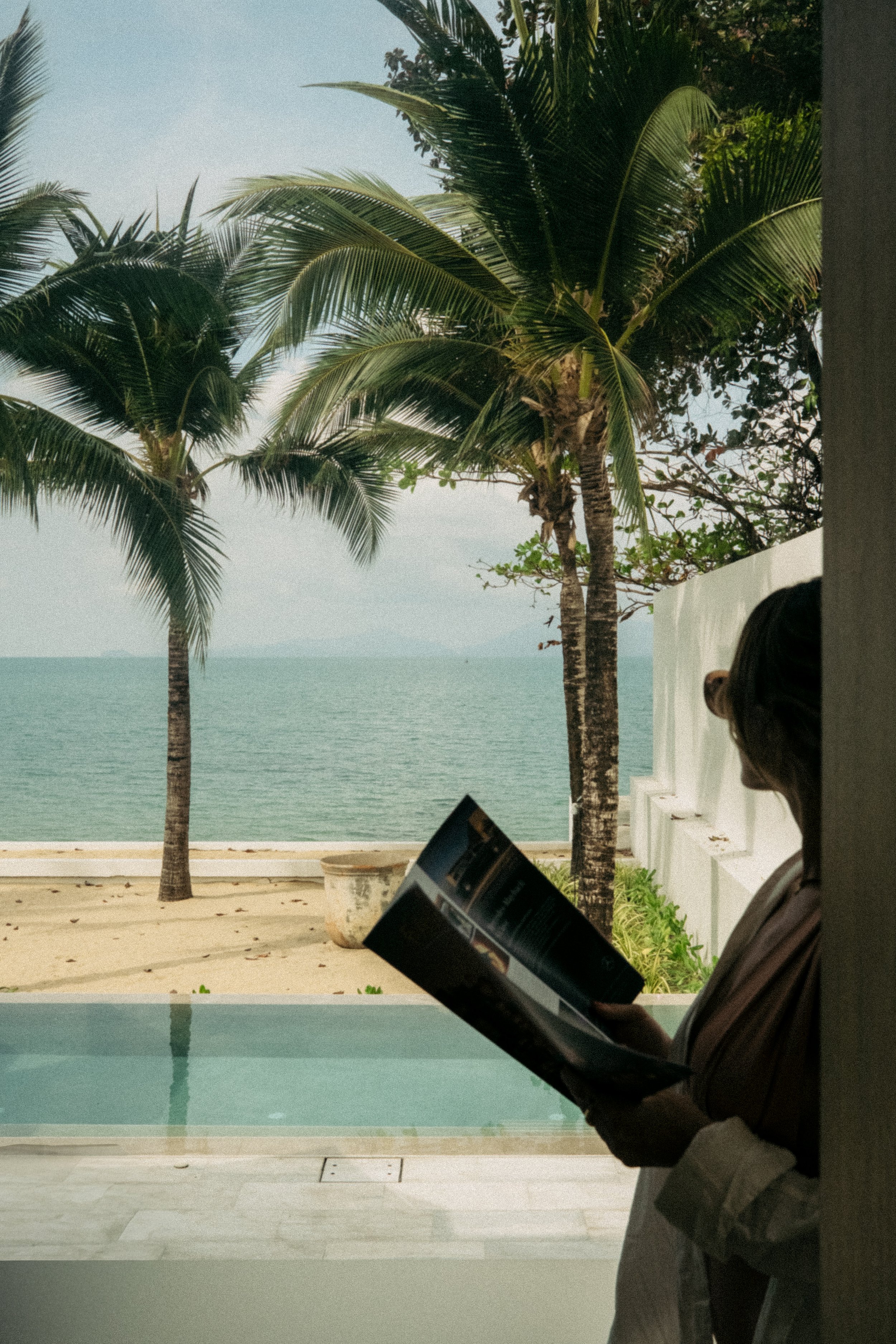 Une femme lit un magazine à l'intérieur avec vue sur des palmiers, la plage et la mer en arrière-plan