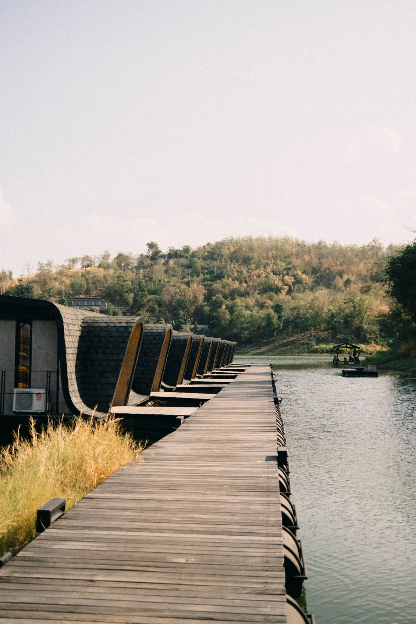 Une passerelle en bois longe un lac avec des bâtiments aux formes arrondies sur le côté gauche et une végétation verdoyante au loin.