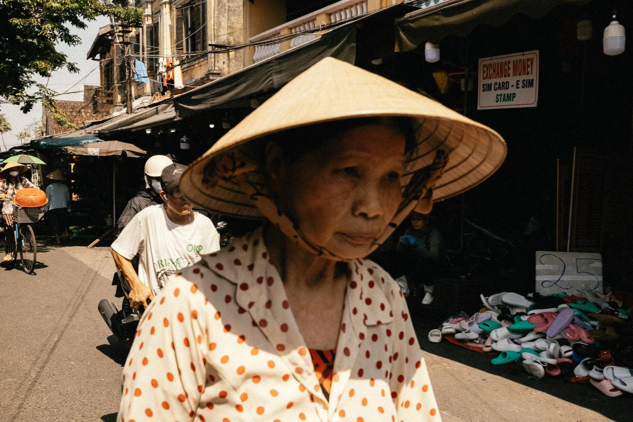 Une femme portant un chapeau conique vietnamien et une chemise à pois, marchant dans une rue animée avec des vendeurs de chaussures et un panneau d'échange de devises en arrière-plan.