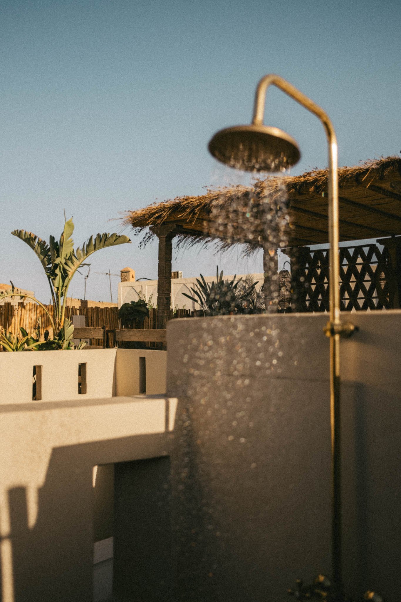 Douche de plein air avec toit en chaume, plantes tropicales, et bâtiments en arrière-plan, sous un ciel clair.
