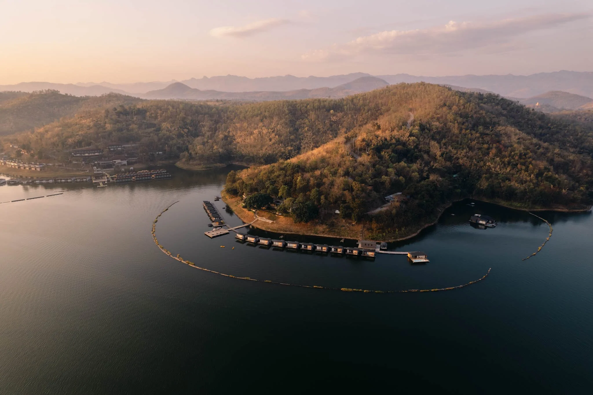 Vue aérienne d'une île arborée entourée d'eau, avec des bungalows sur l'eau et des montagnes à l'arrière-plan au coucher du soleil.