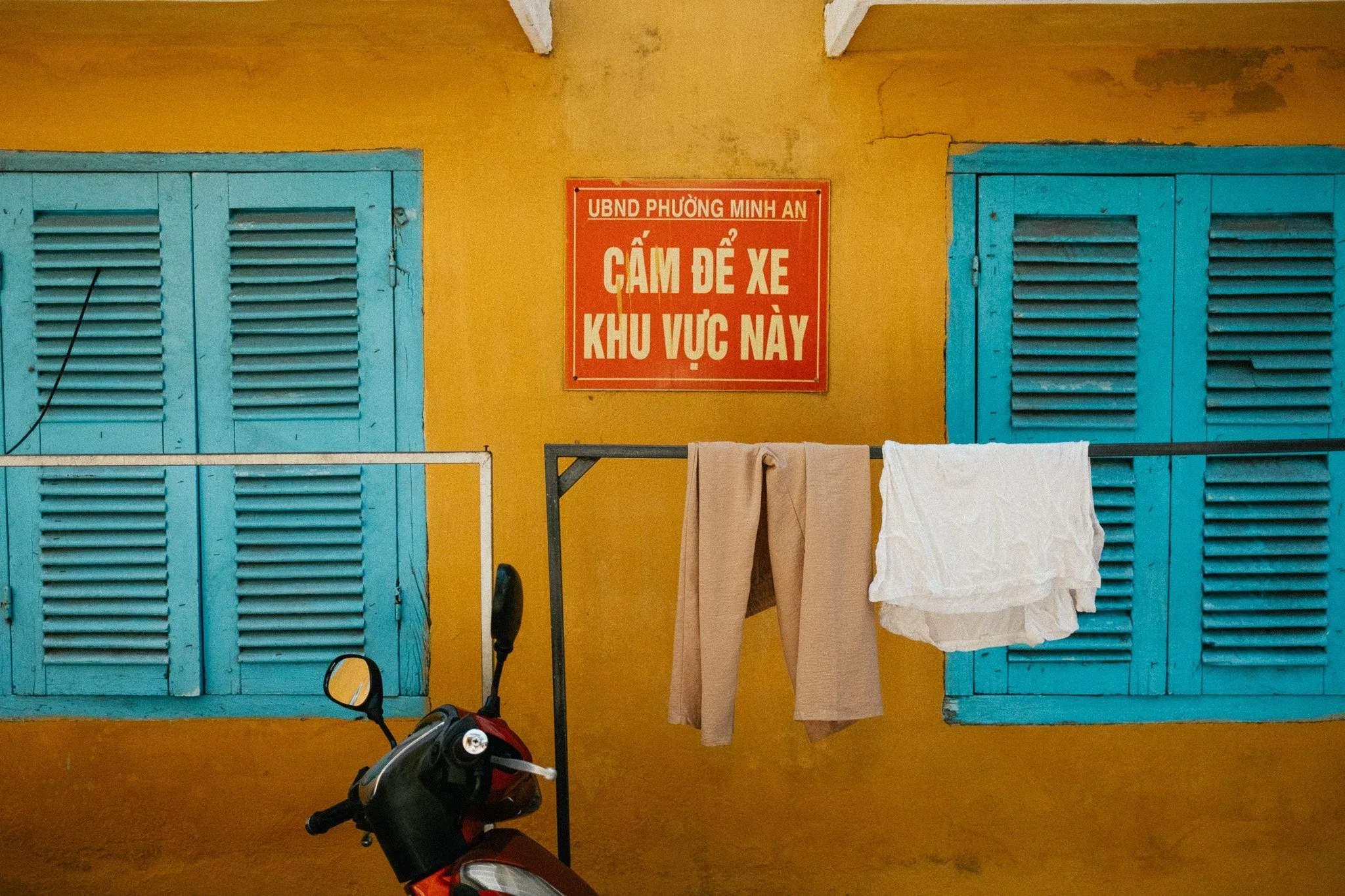 Panneau rouge avec texte en vietnamien, fenêtres bleues à volets, linge suspendu et un scooter dans une maison jaune