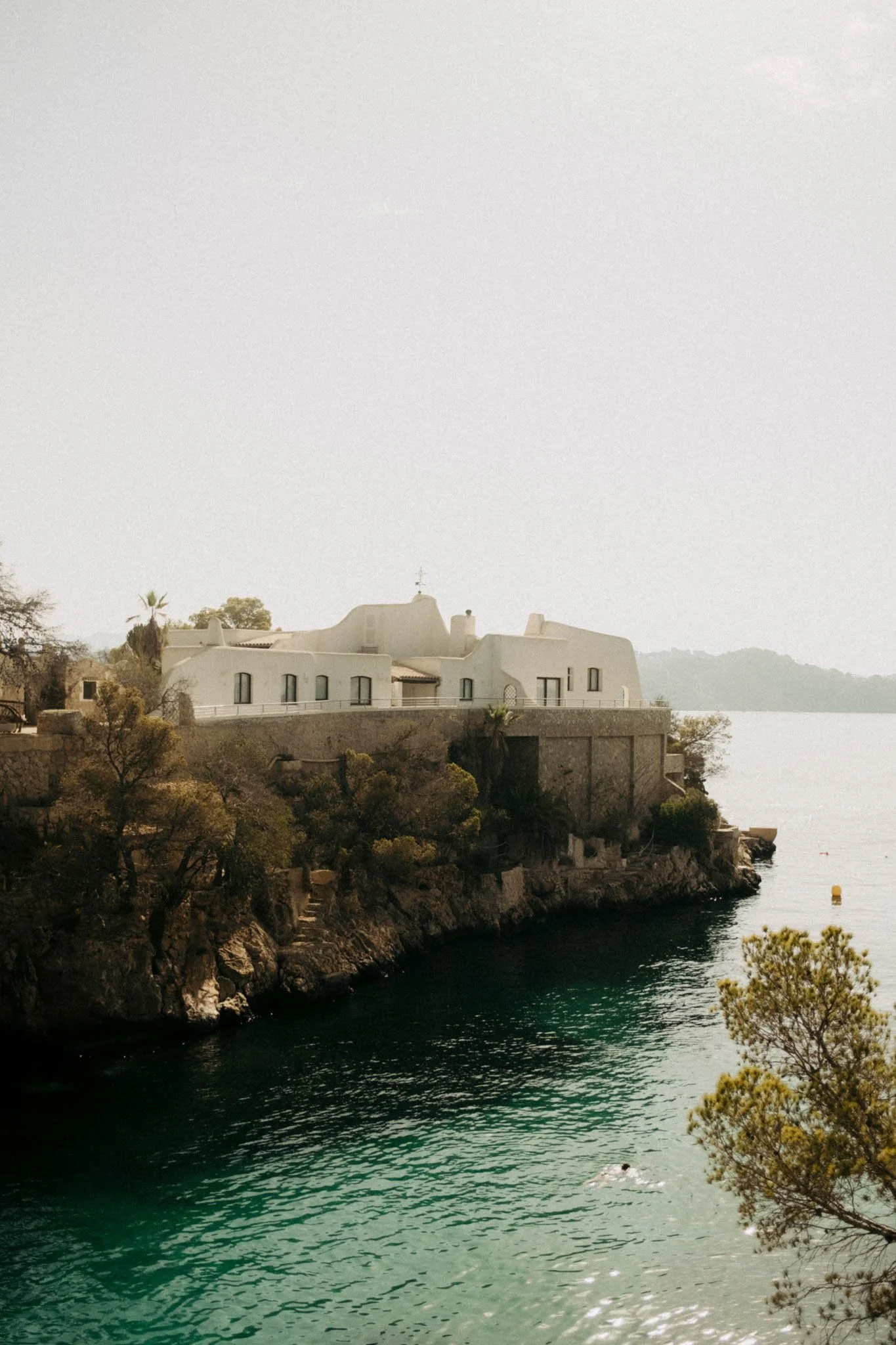 Une maison blanche perchée sur une falaise rocheuse au bord de l'eau, avec quelques arbres et un if en premier plan.
