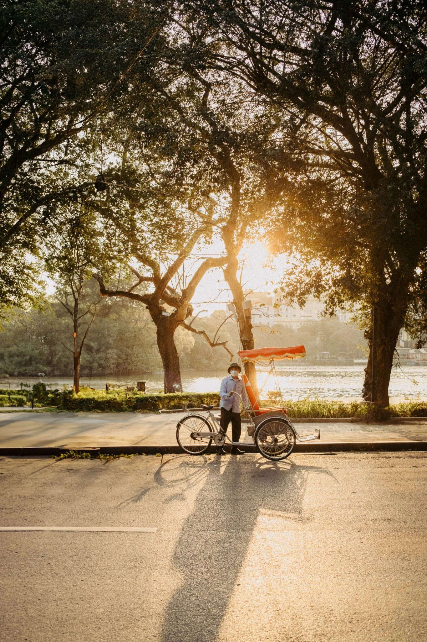 Un homme avec un masque sanitaire, portant une chemise bleue, se tient près d'une pousse-cousse orange sur le trottoir au lever du soleil avec des arbres au fond.