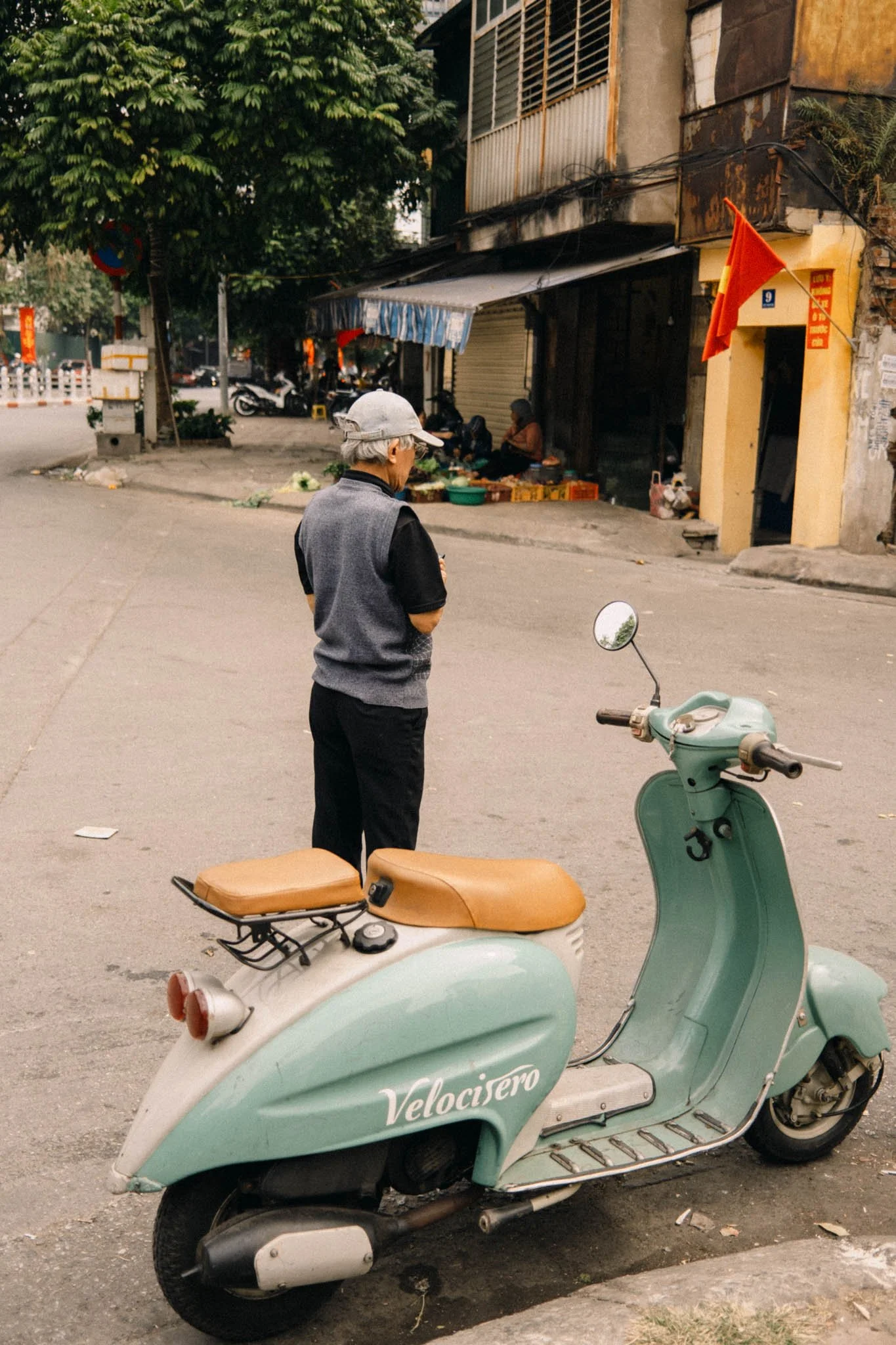 Une personne âgée regardant une moto de type scooter bleu pastel, avec une boutique en arrière-plan et des arbres.