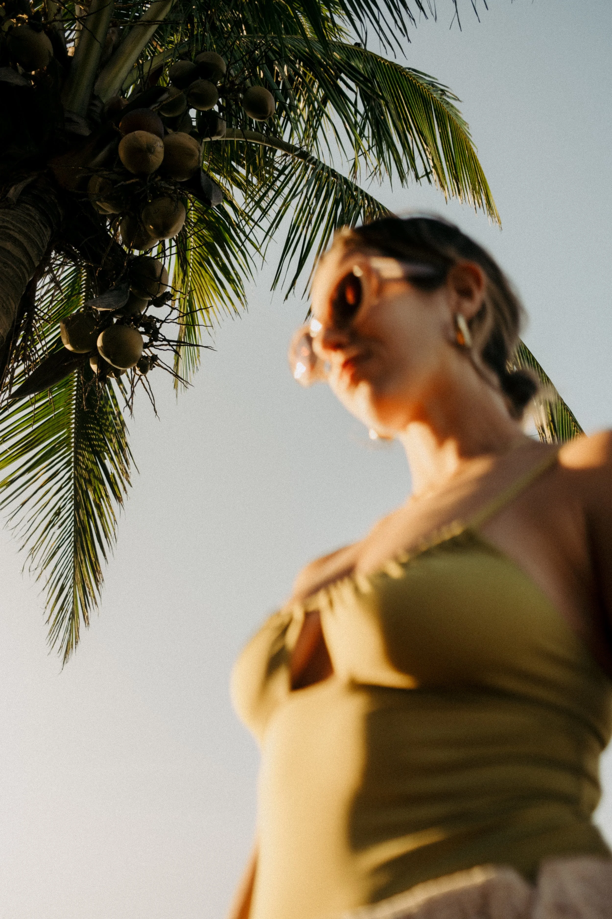 Une femme portant des lunettes de soleil et une robe beige, vue de dessous contre un arbre de noix de coco et un ciel clair.