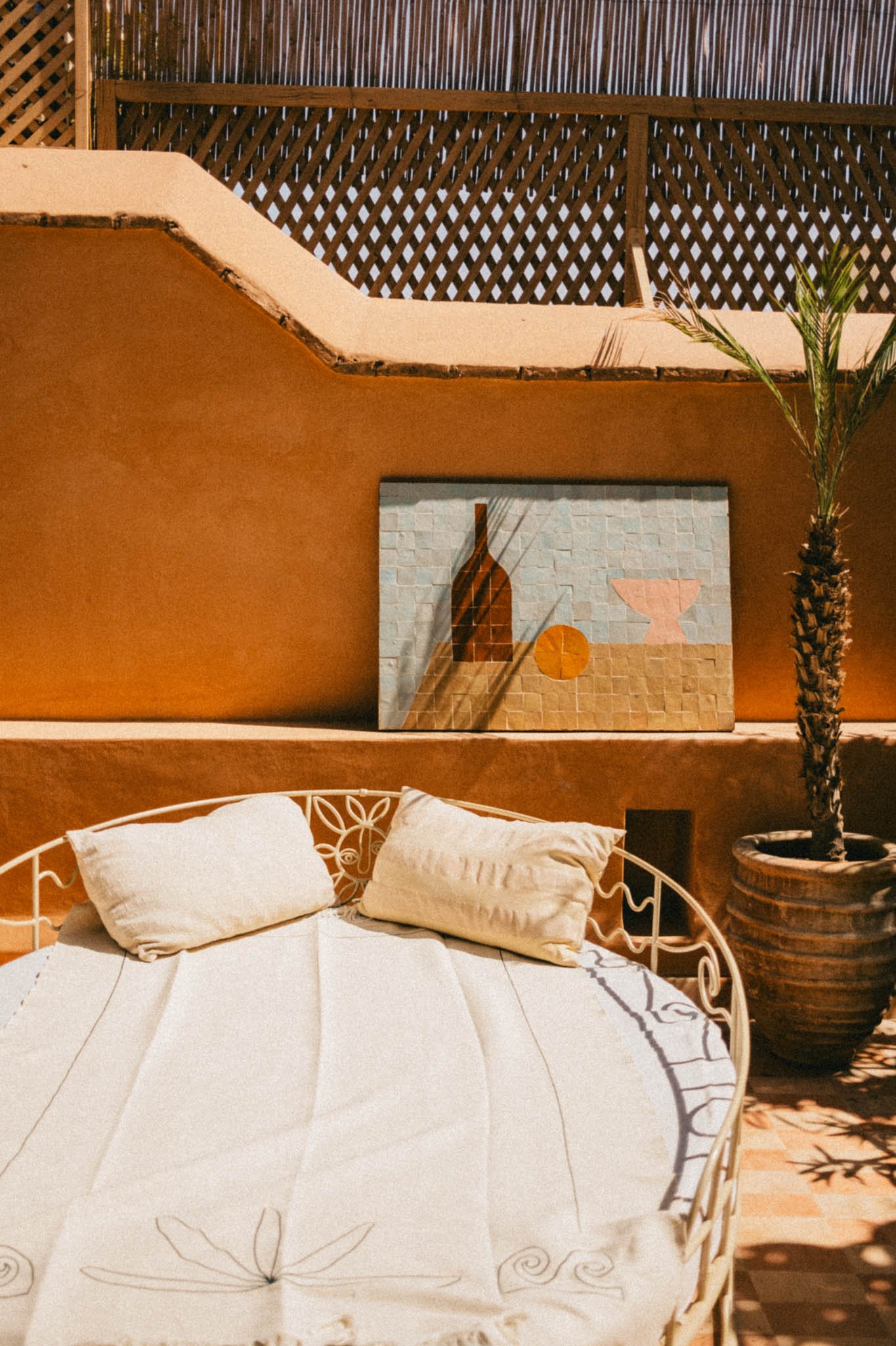 Une terrasse décorée avec un lit en fer forgé et deux coussins, une peinture abstraite sur le mur en arrière-plan, et un palmier dans un pot en terre cuite. L'éclairage chaud crée une ambiance chaleureuse.