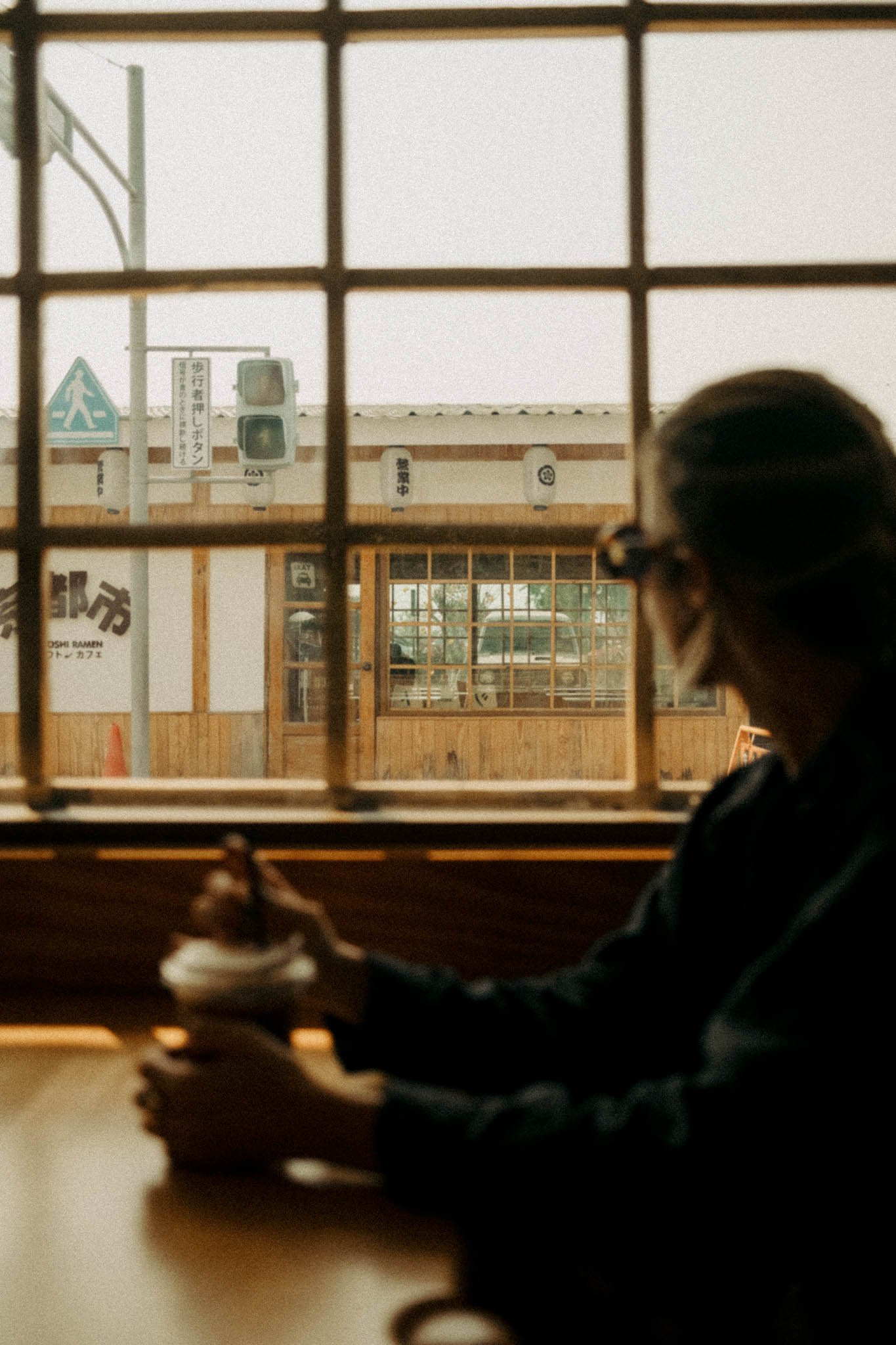 Une femme avec des lunettes regarde par une fenêtre en bois, l'extérieur montre un bâtiment en bois avec des panneaux japonais, des lampes en papier et une lumière naturelle douce.