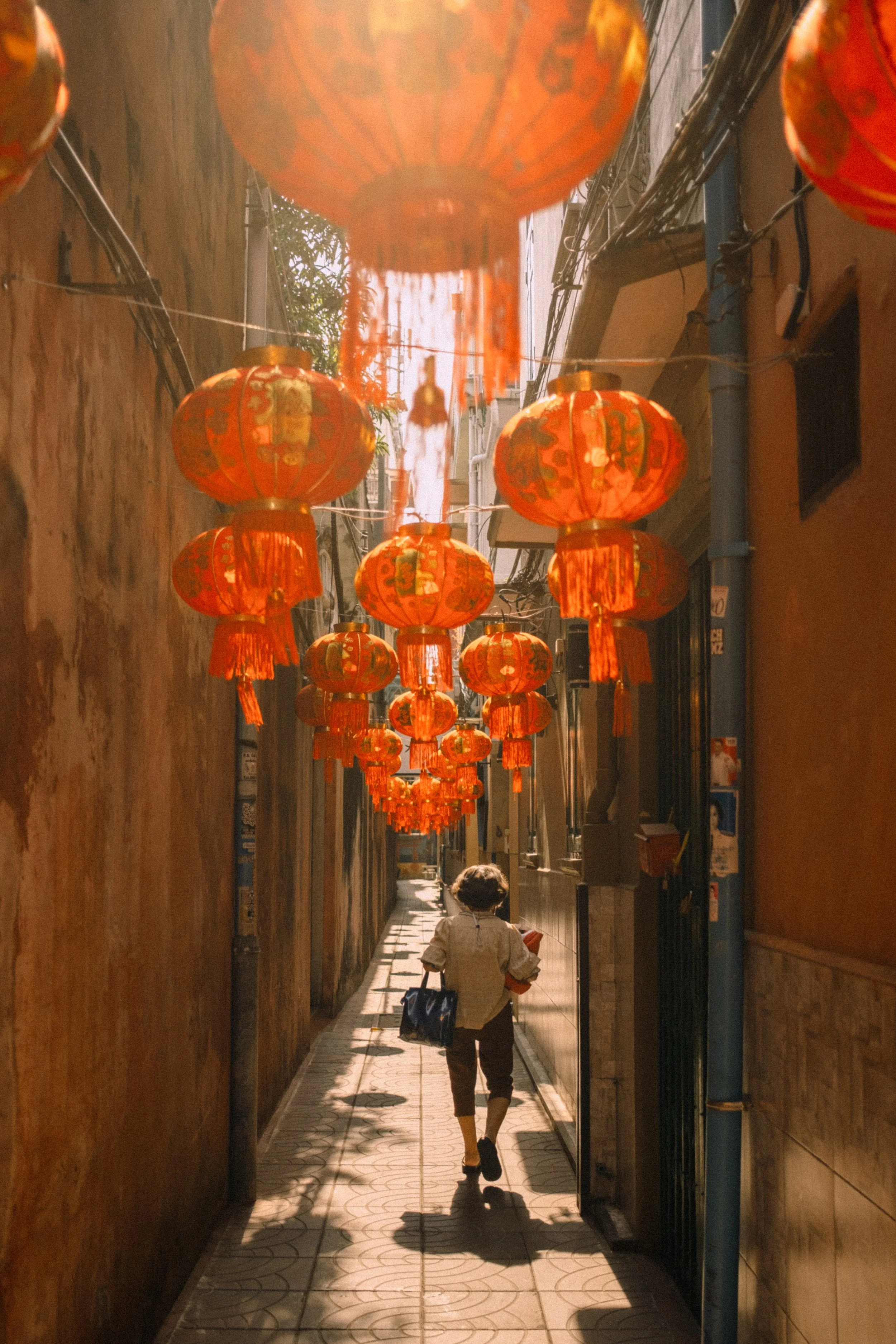 Une petite fille marche dans une ruelle étroite décorée de lanternes chinoises orange suspendues au plafond, avec des murs en pierre et des fils électriques au-dessus.