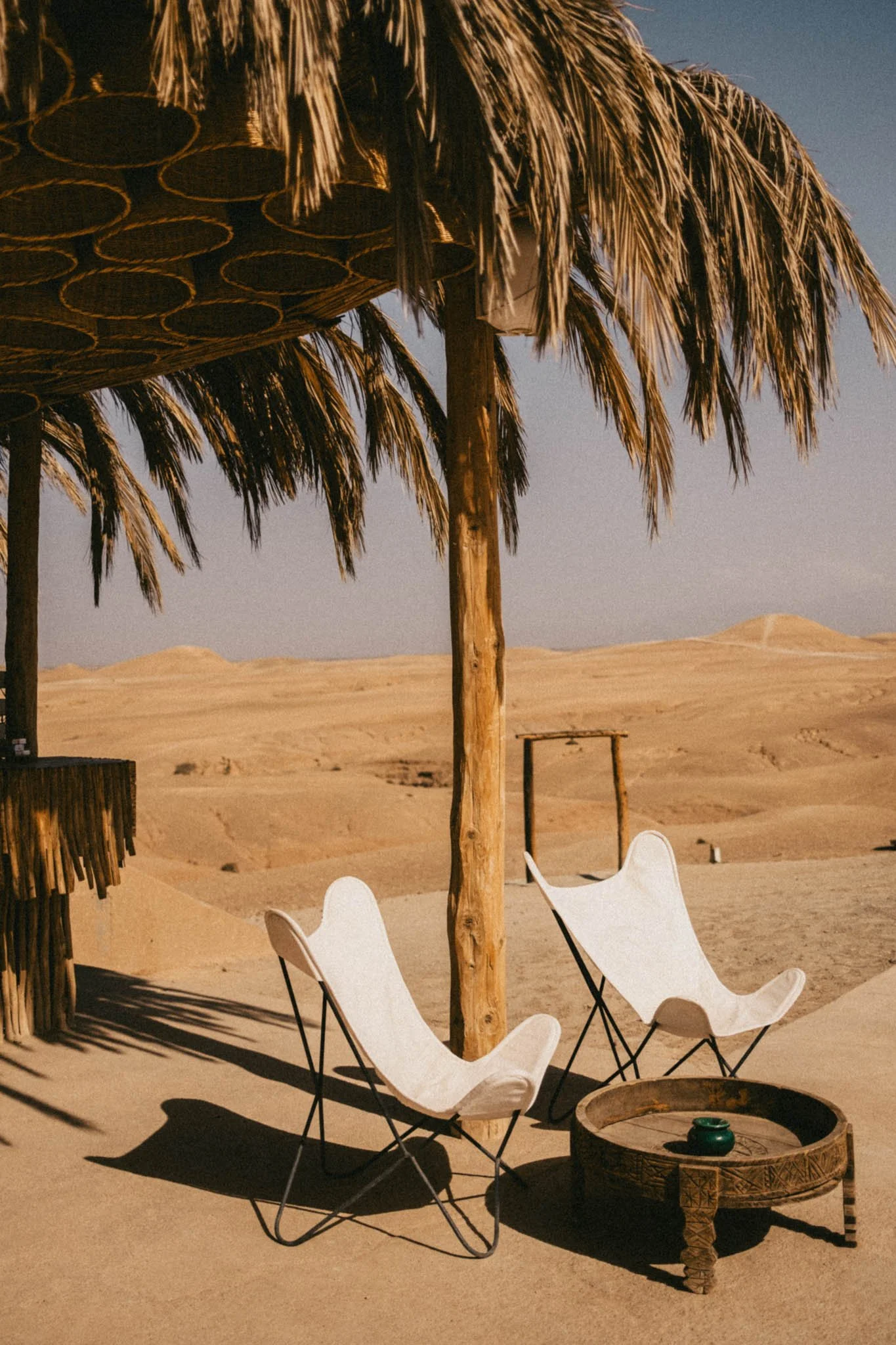 Chaises en tissu blanc sous un parasol en feuilles de palmier dans un désert avec dunes de sable.