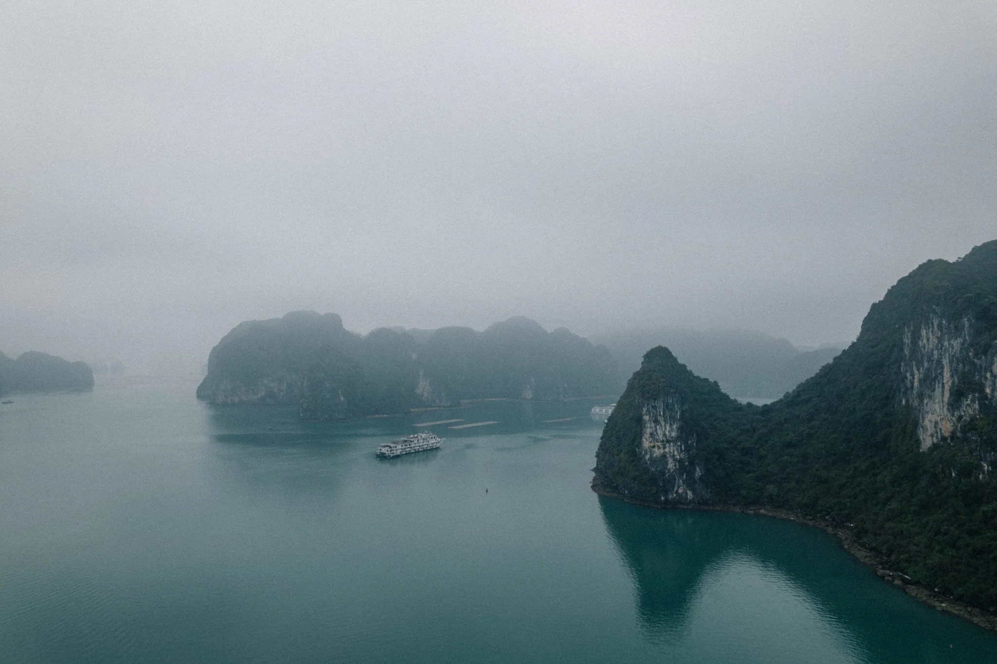 Vue aérienne d'une baie avec des montagnes karstiques recouvertes de végétation et deux bateaux naviguant sur l'eau calme, dans un paysage brumeux.
