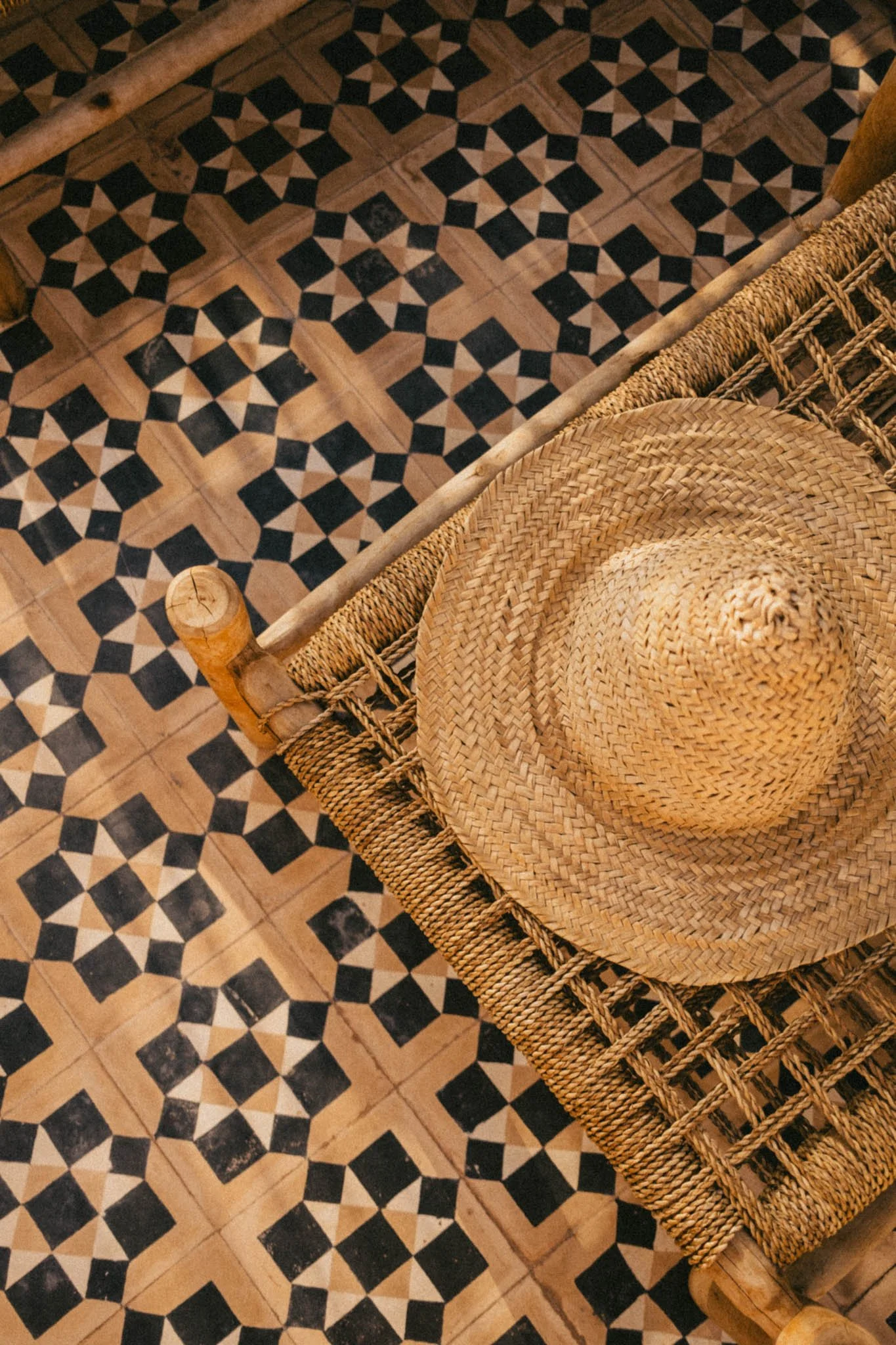 Un chapeau en paille posé sur une chaise en rotin, vue du dessus, avec un sol en carreaux à motif géométrique en noir, beige et marron.