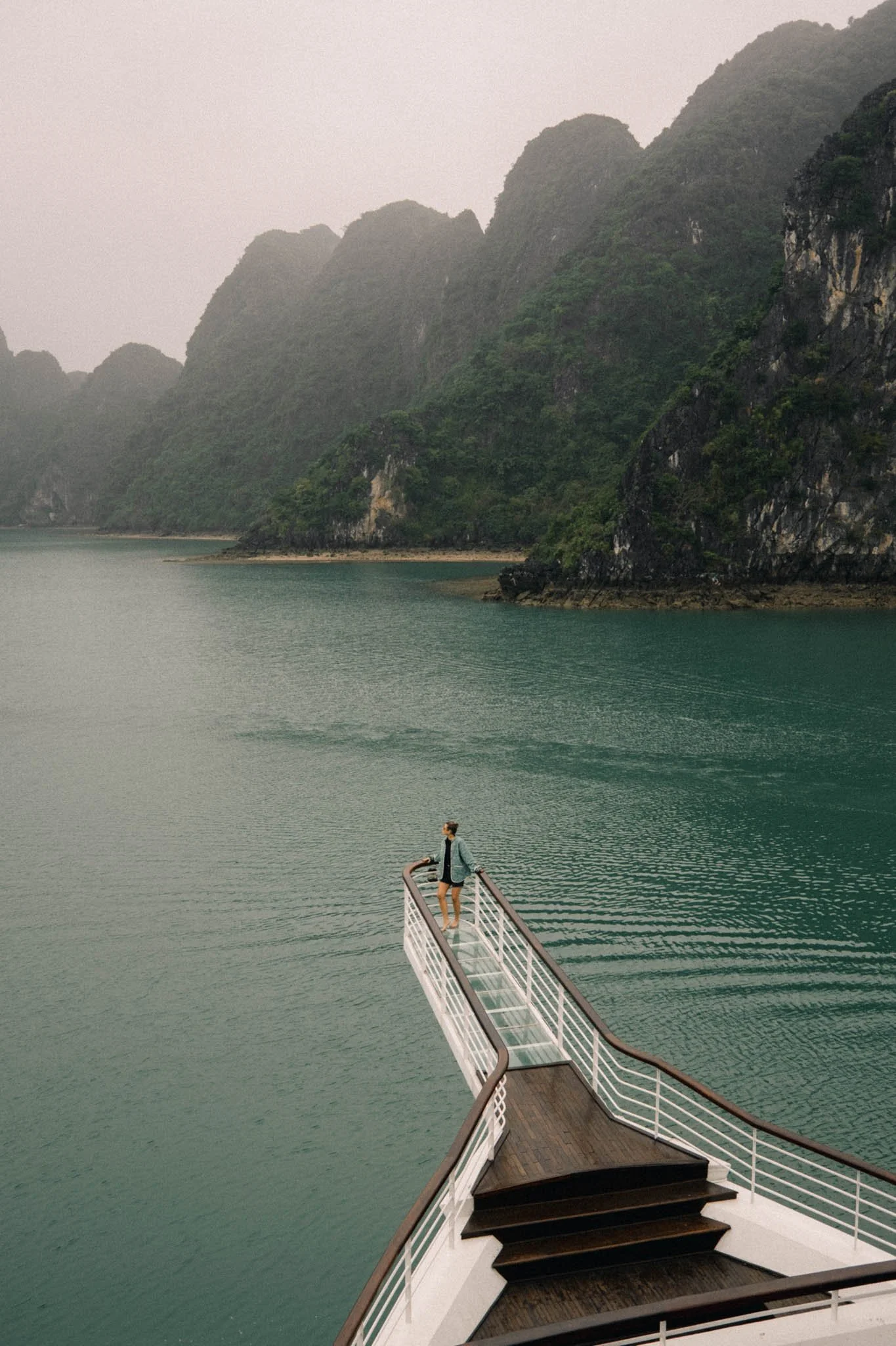 Une femme debout à l'extrémité d'un pont en bois avec une rambarde en métal, s'étendant au-dessus d'une grande étendue d'eau entourée de montagnes verdoyantes, sous un ciel nuageux.