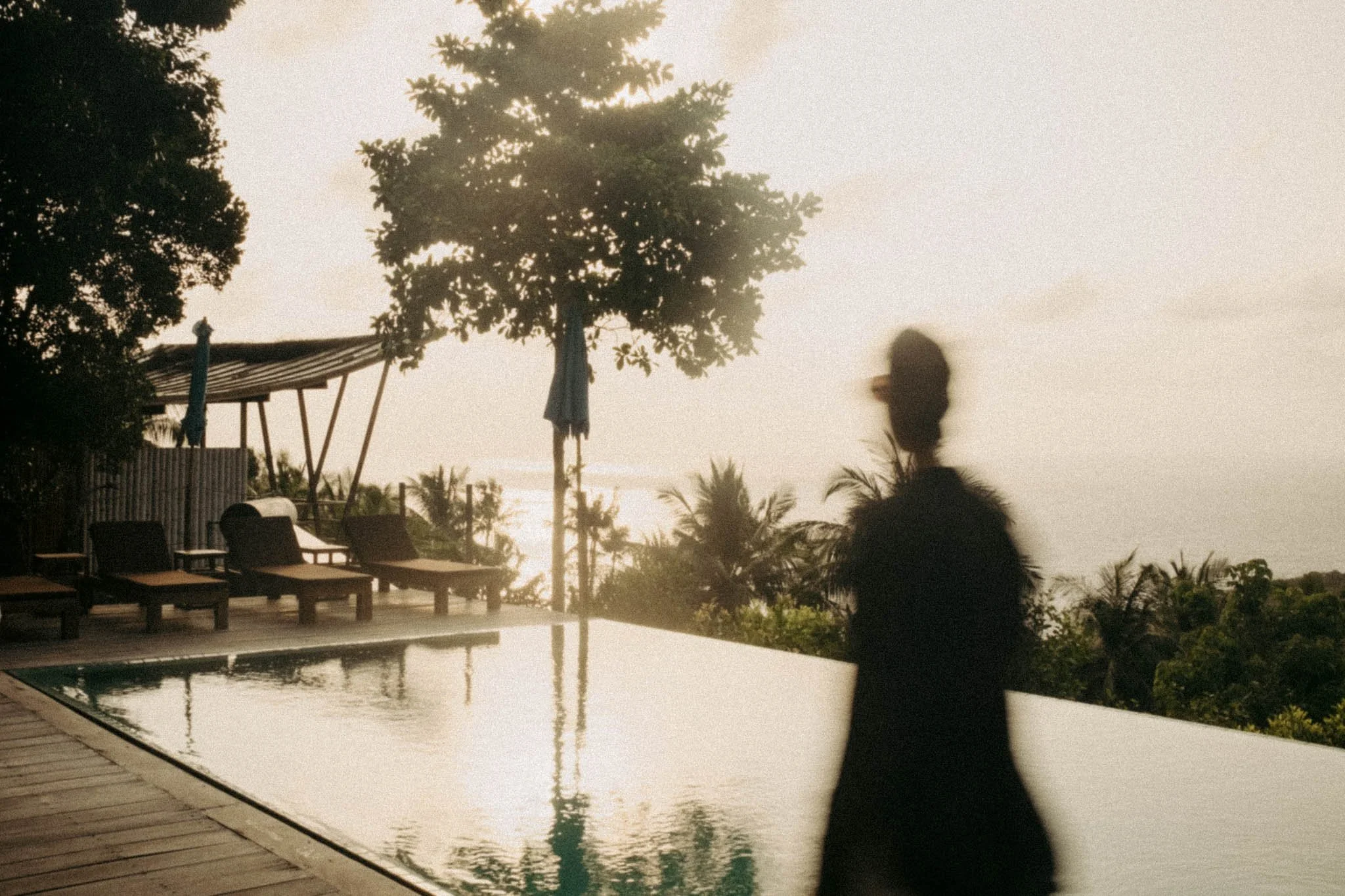 Une silhouette floue d'une personne près d'une piscine lors d'un coucher de soleil, avec des chaises longues, un parasol et des arbres en arrière-plan.