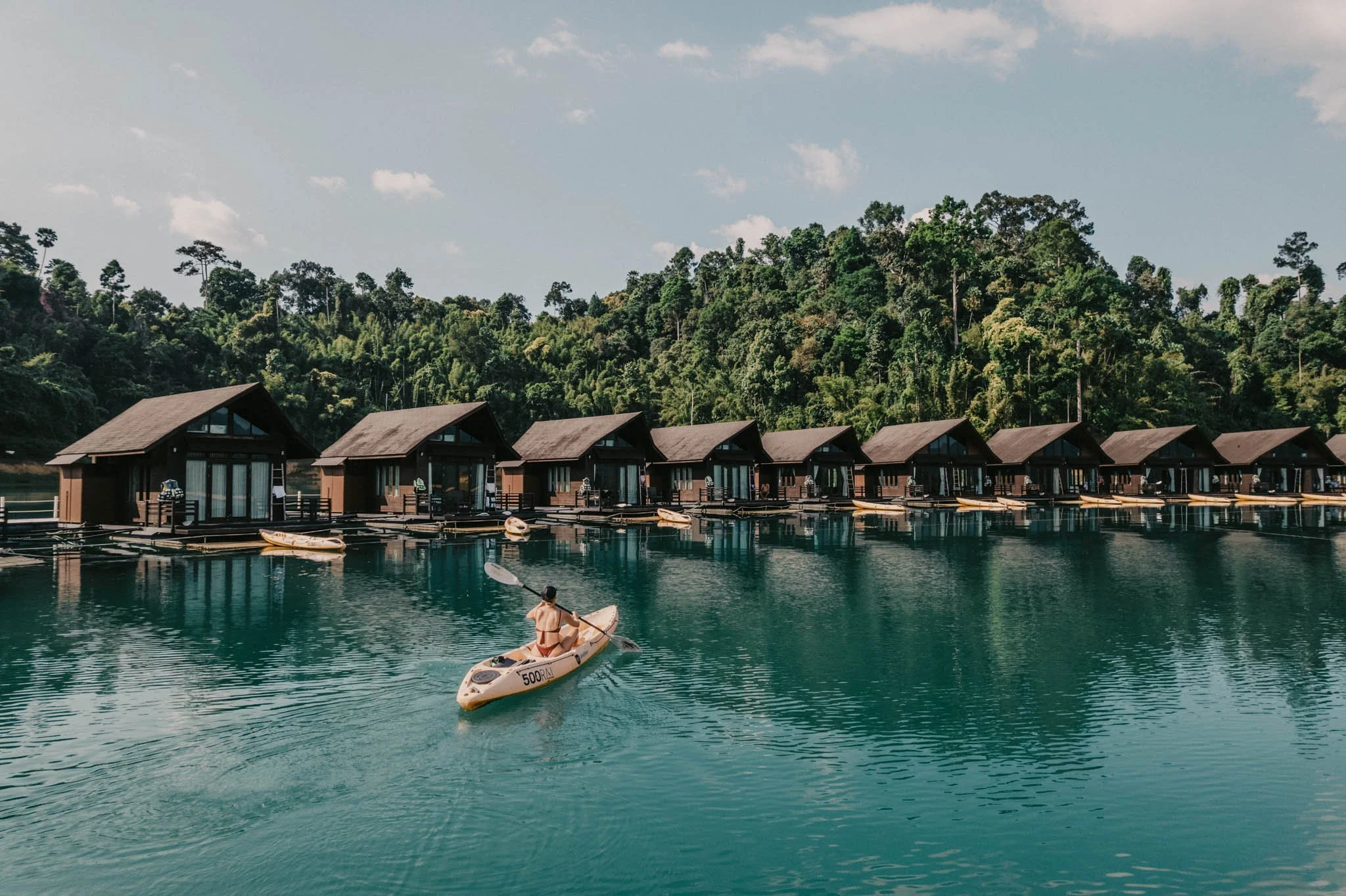 Cabanes en bois au bord d'un lac avec un kayakiste âgé, entouré de forêt dense et ciel partiellement nuageux.