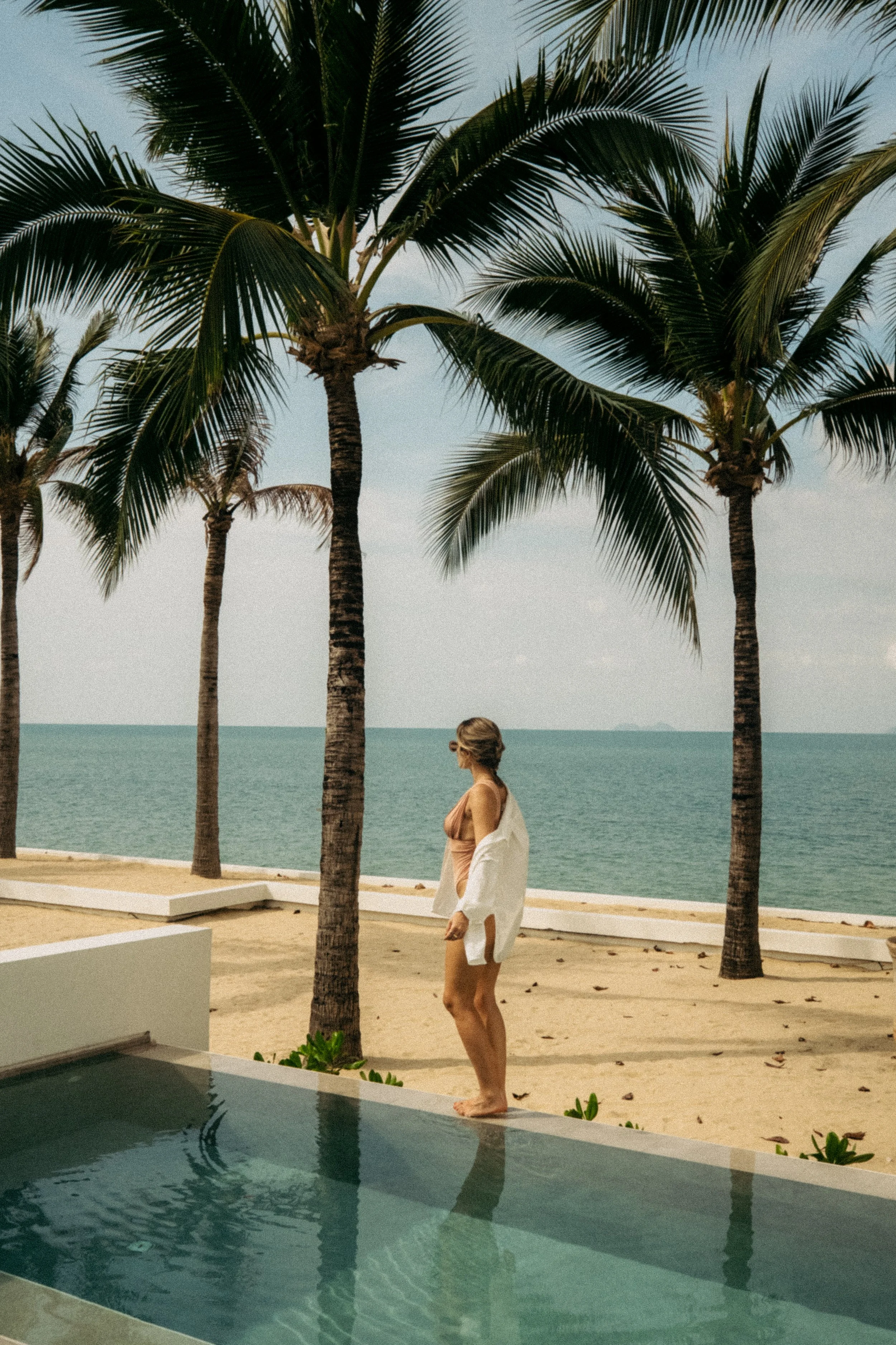Une femme en maillot de bain et chemise blanche regarde vers la mer, debout au bord d'une piscine, avec des palmiers en arrière-plan sur une plage ensoleillée.