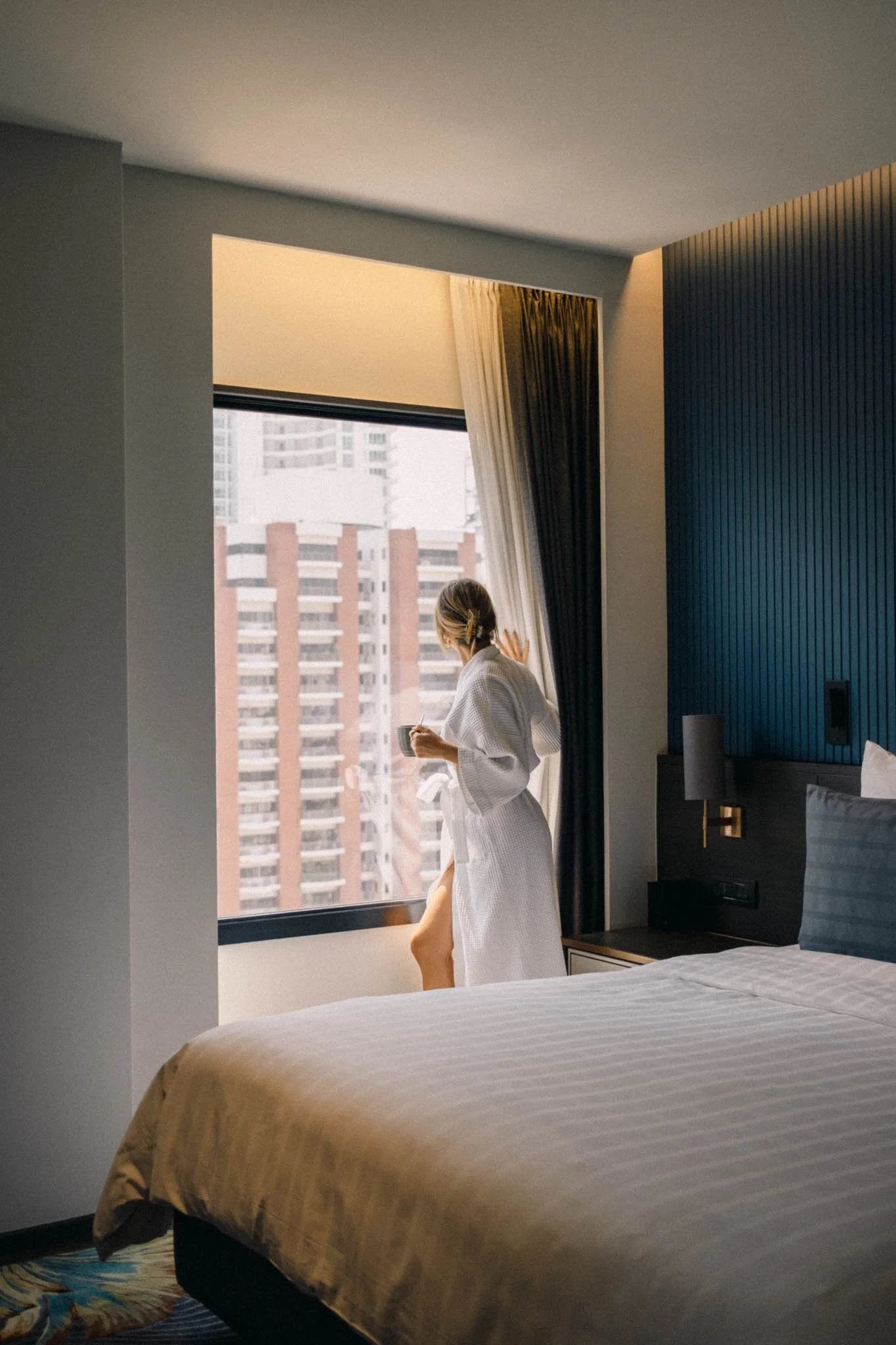 Femme en peignoir blanc regardant par la fenêtre dans une chambre d'hôtel moderne.