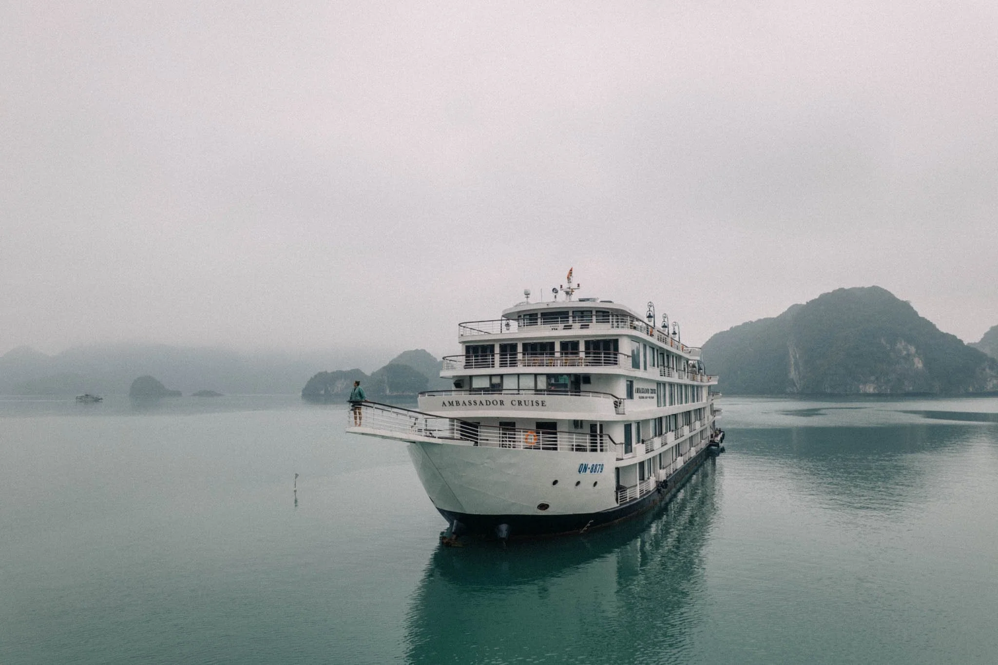 Un bateau de croisière blanc nommé 'Ambassador Cruise' flotte sur une mer calme avec des îles rocheuses en arrière-plan, dans un ciel brumeux.
