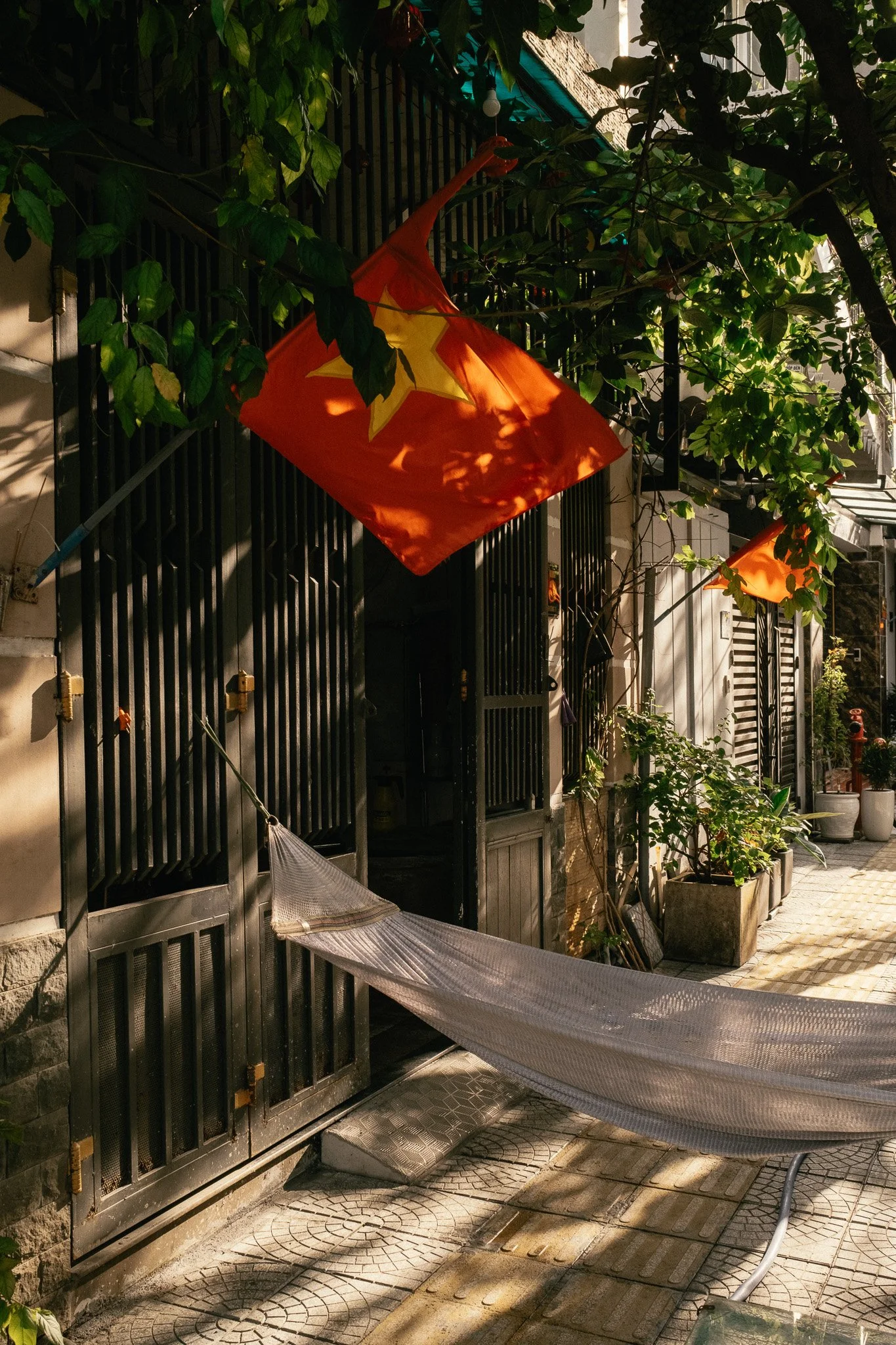 Balcon avec un drapeau rouge à étoile jaune, hamac, plantes en pots, et lumière du soleil créant des ombres